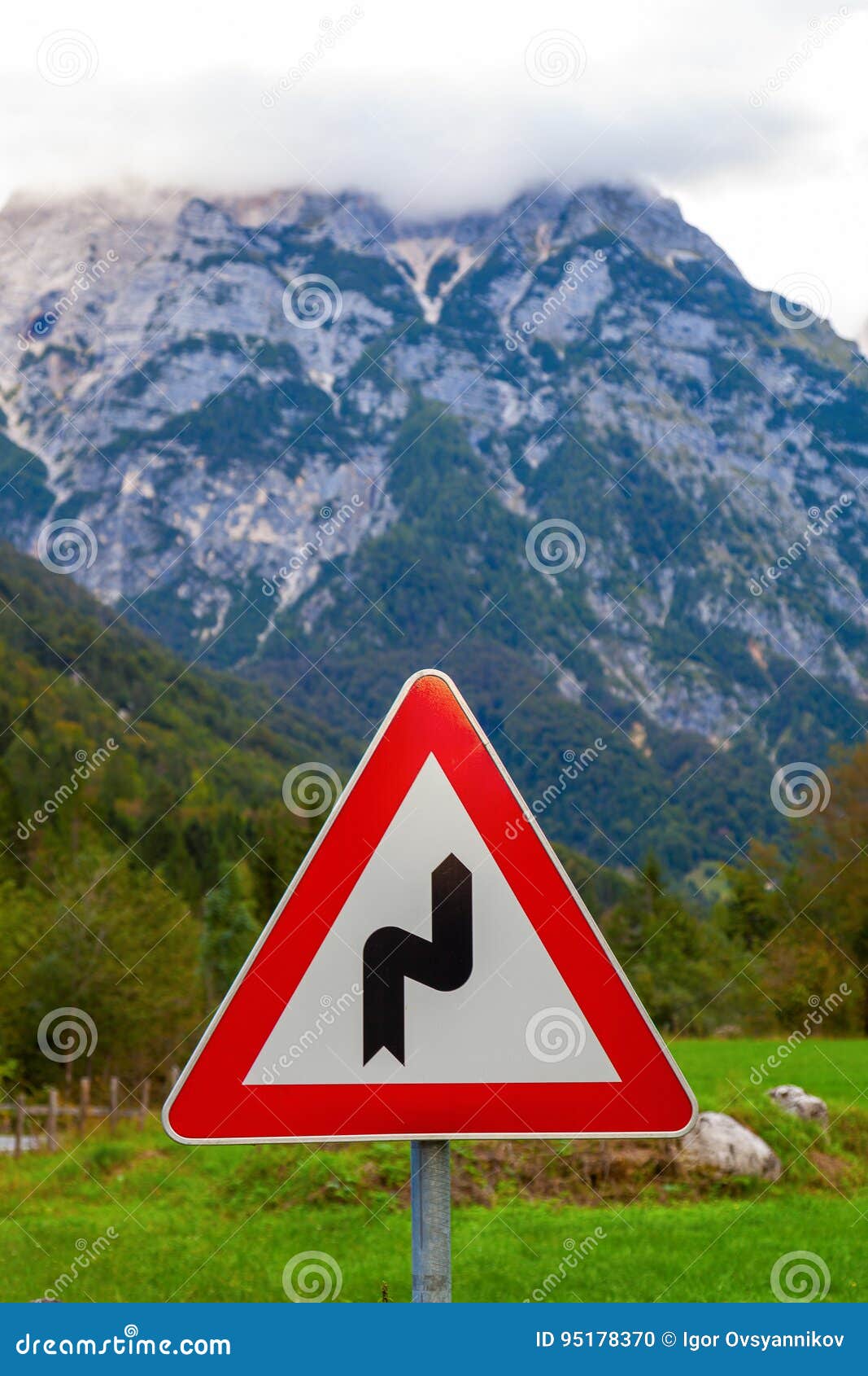 Señal de tráfico en Julian Alps, Eslovenia. ` Peligroso de la vuelta del ` de la señal de tráfico en un fondo borroso de la hierba verde y de las montañas en las nubes