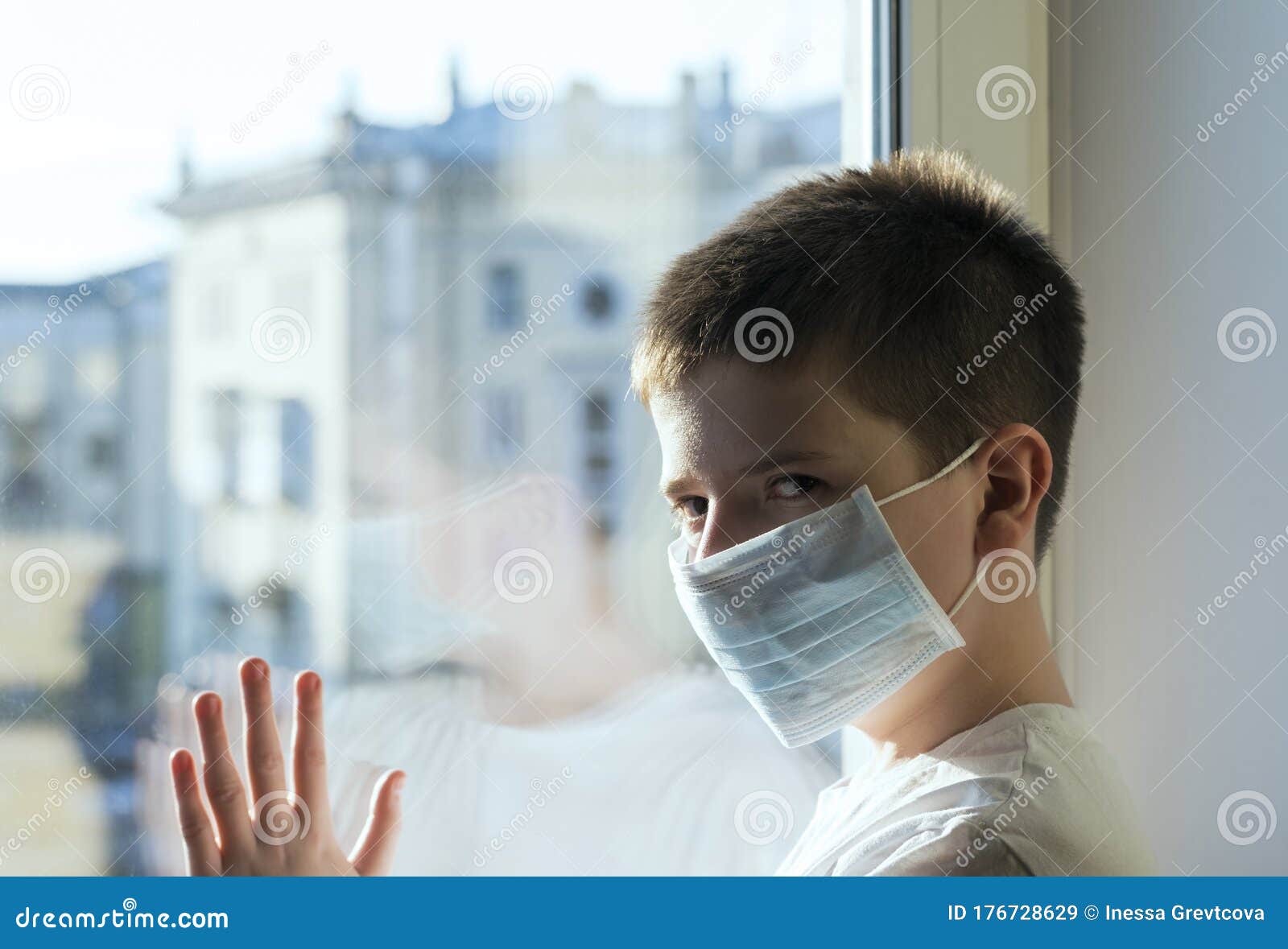 a sick boy in a medical mask stands at the window and looks longingly.