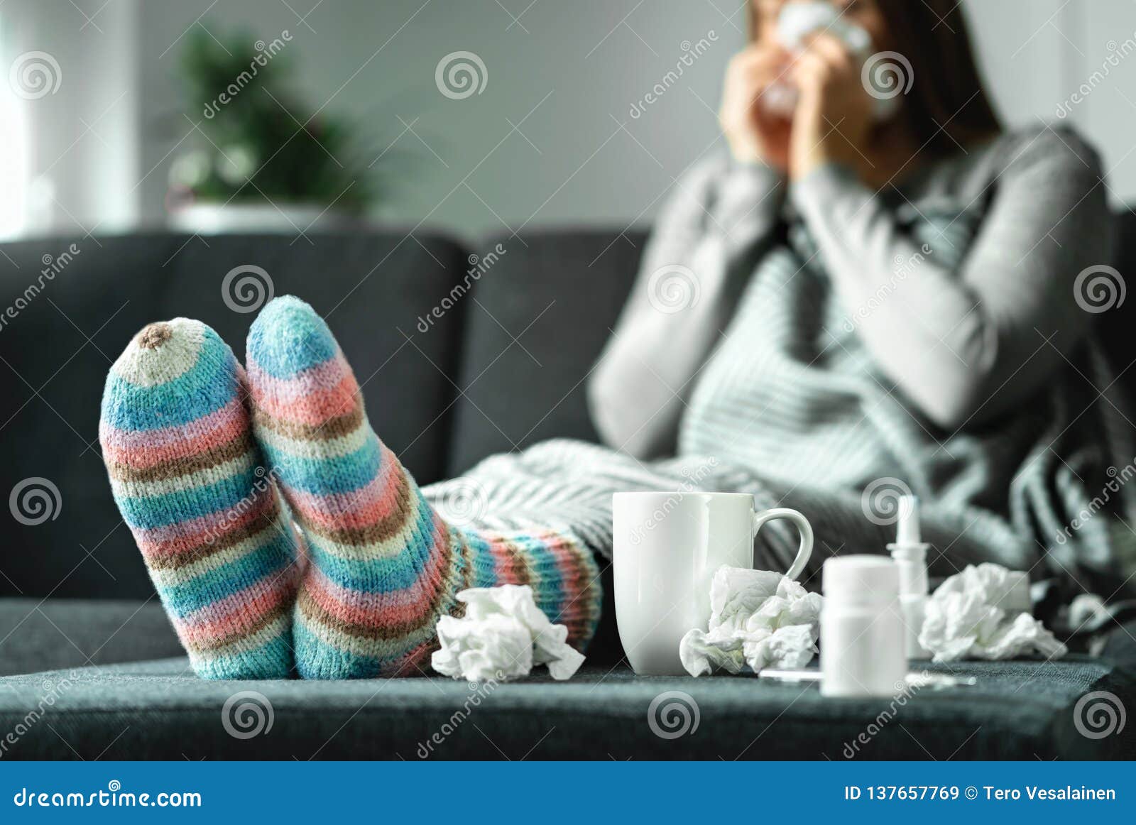 sick woman with flu, cold, fever and cough sitting on couch at home. ill person blowing nose and sneezing with tissue.