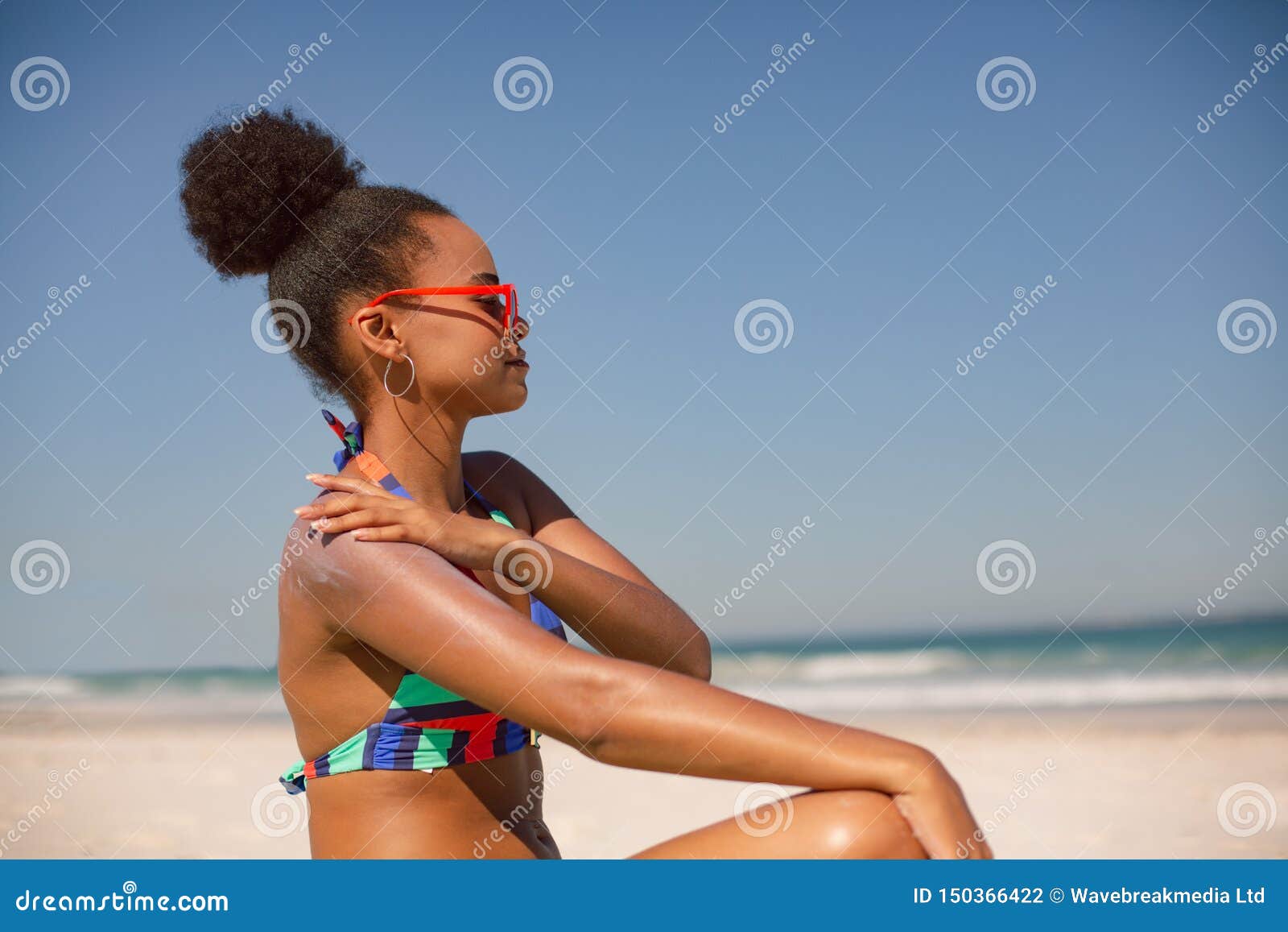 woman in bikini applying sunscreen lotion on shoulder at beach in the sunshine