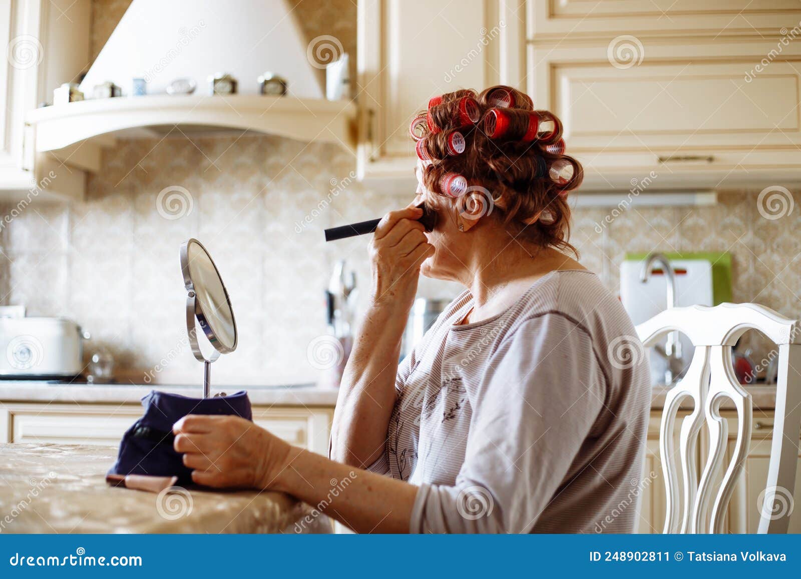 side view of elderly woman in pink hair-curlers sitting at table, looking at mirror, putting powder rouge on skin.