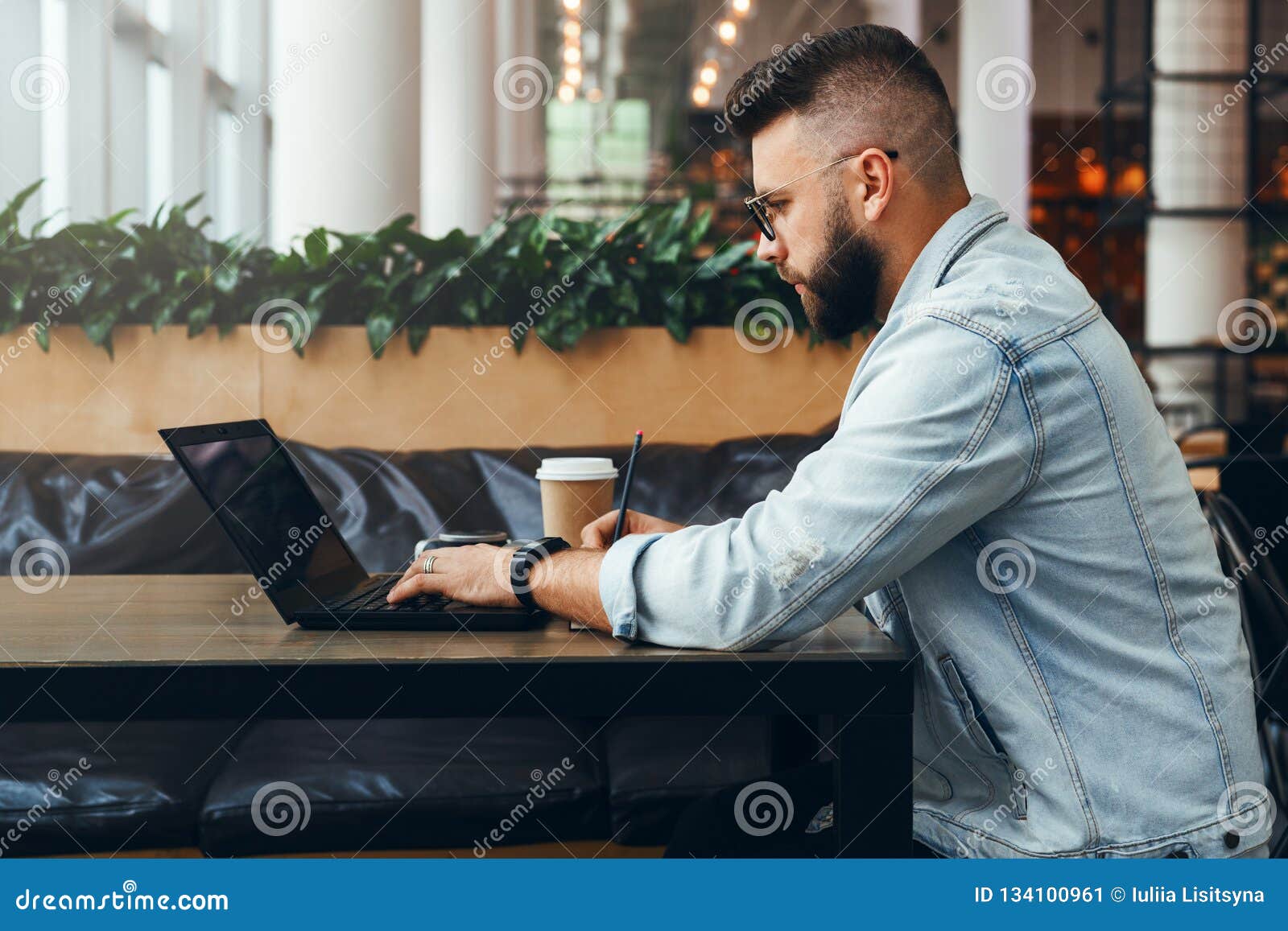 side view.hipster businessman sitting indoors,writing notes in notepad and working on laptop for solving working issues