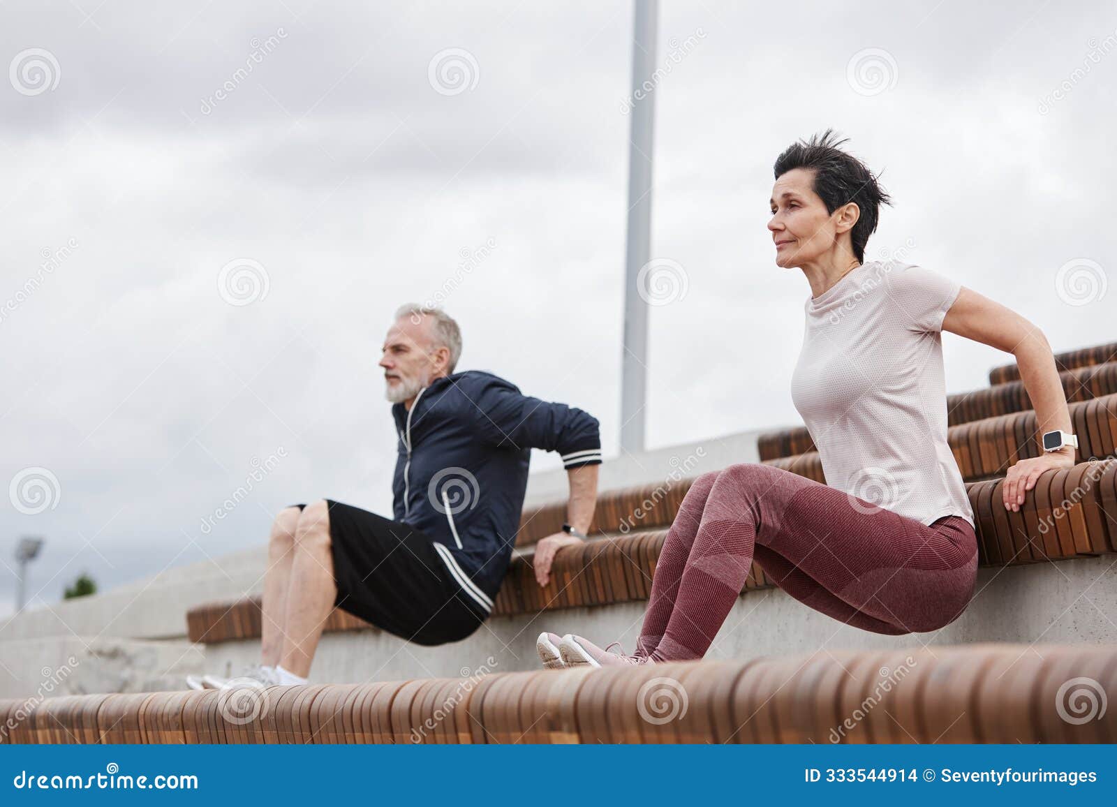 elderly couple doing tricep bench dips on workout at park against gray sky