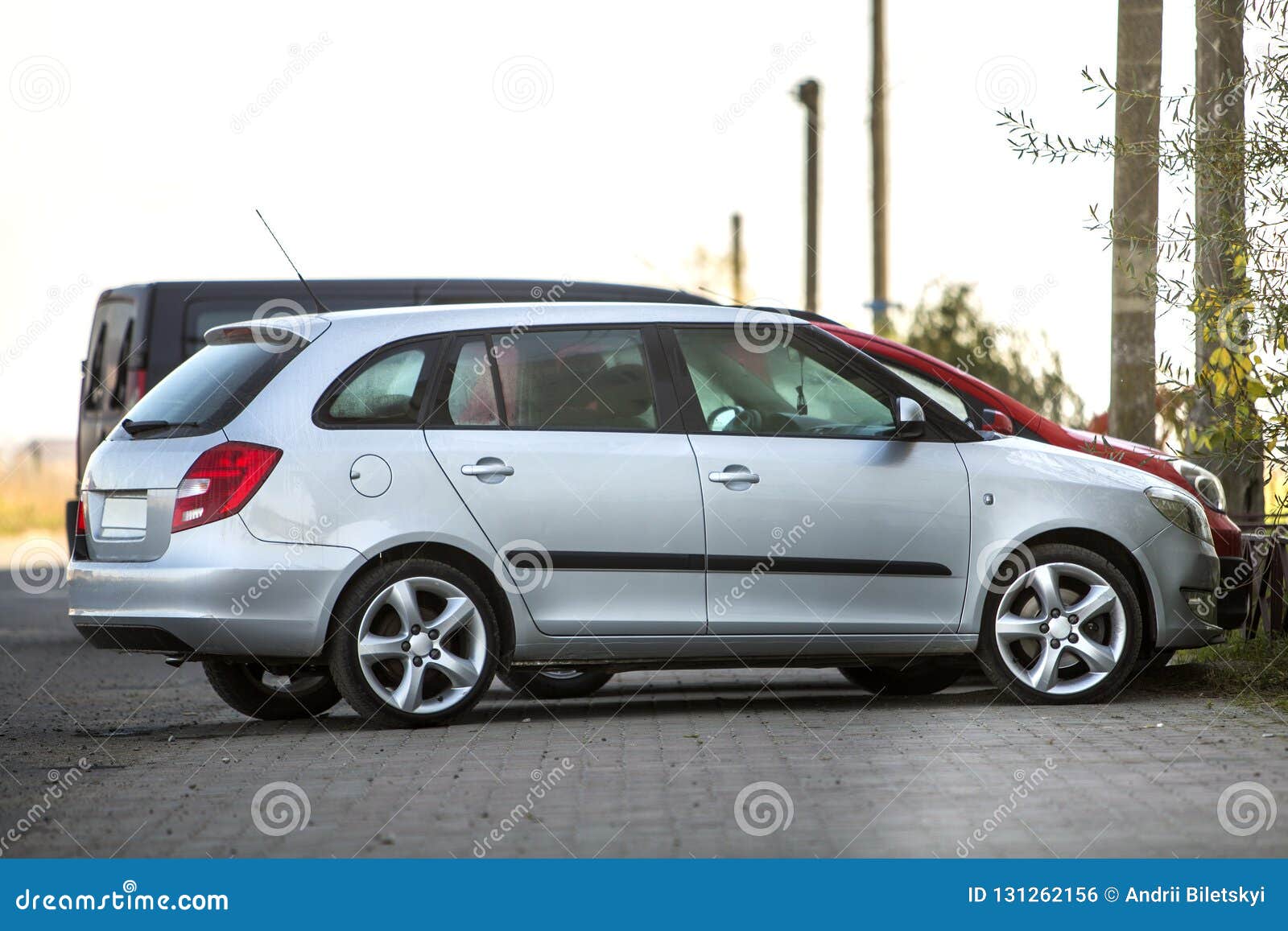 side view of silver shiny luxurious car parked on empty paved road at low iron fence on background of small red mini vehicle and
