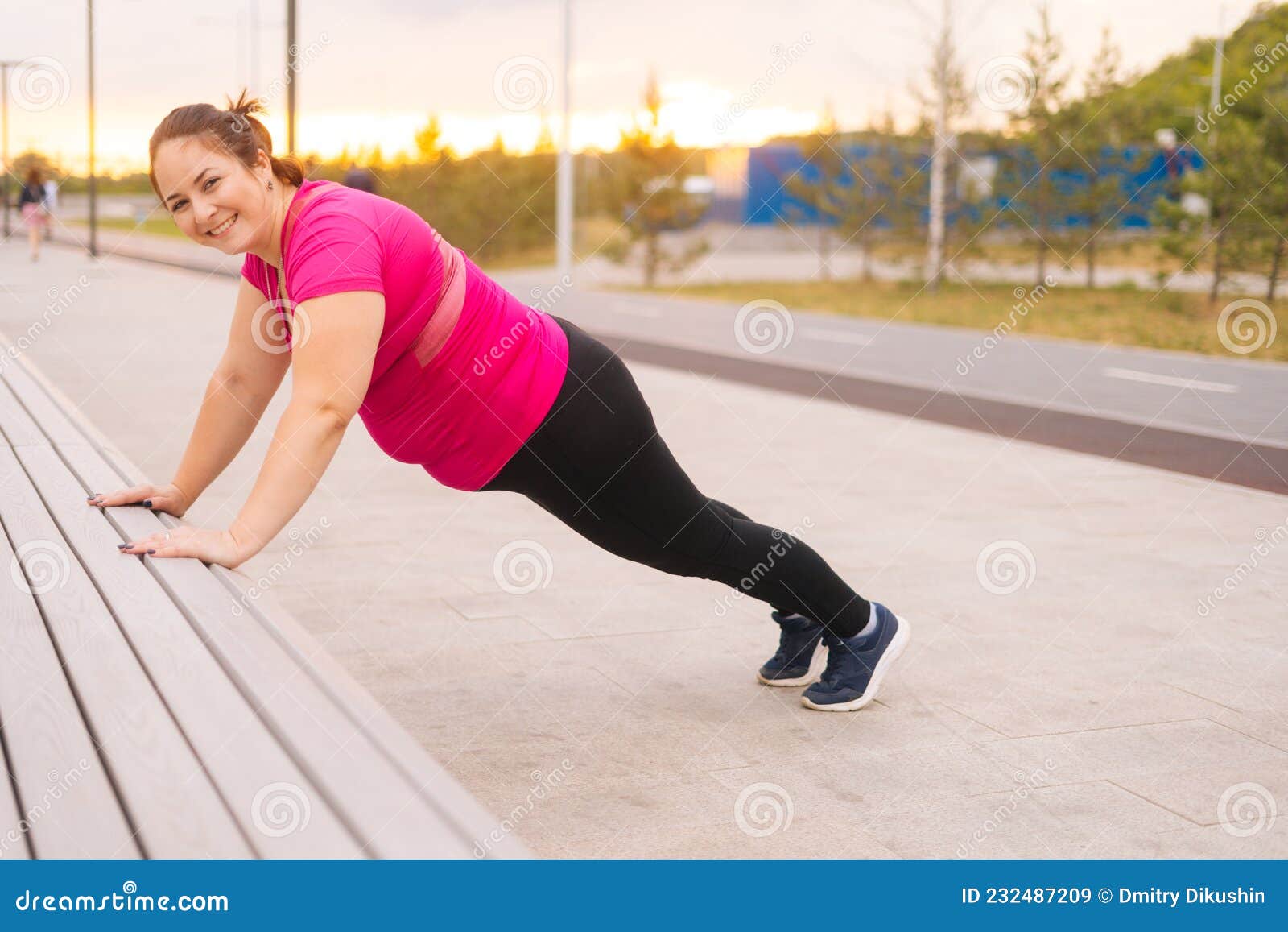 Side view of strong mature redhead woman in leggings doing push-ups from  bench in city park Stock Photo