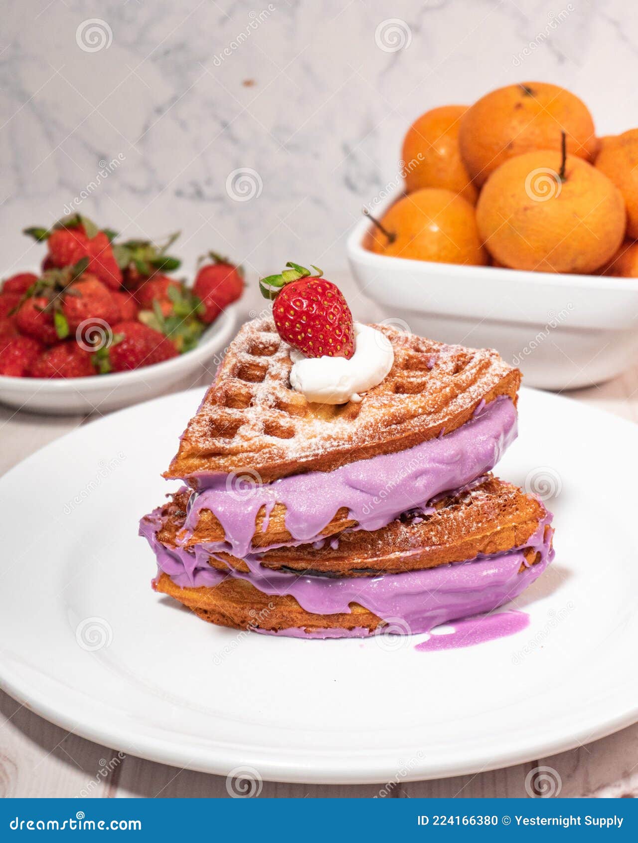 side view of a waff,e with purple cream and  a strawberry topping. with oranges and strawberries in the background