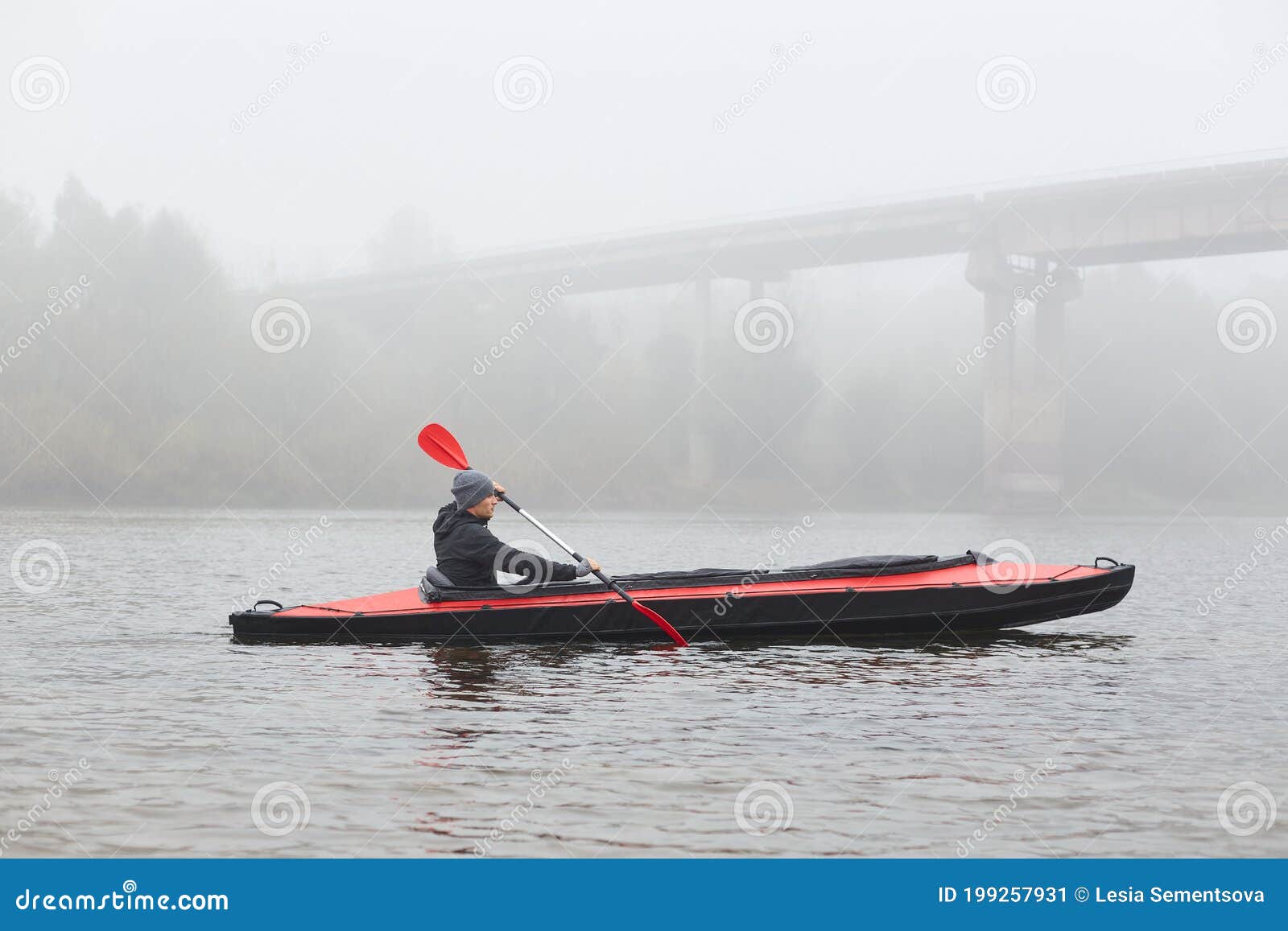 side view of young kayaker in river, padding, man enjoying water sport in foggy morning with bridge on background, male rowing