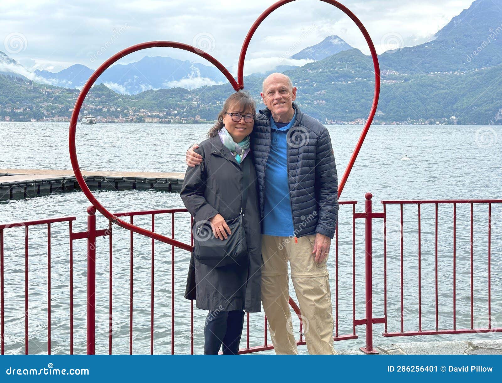 sixty-five year-old korean tourist and her seventy-six year old caucasian husband posing in a a heart beside lake como.