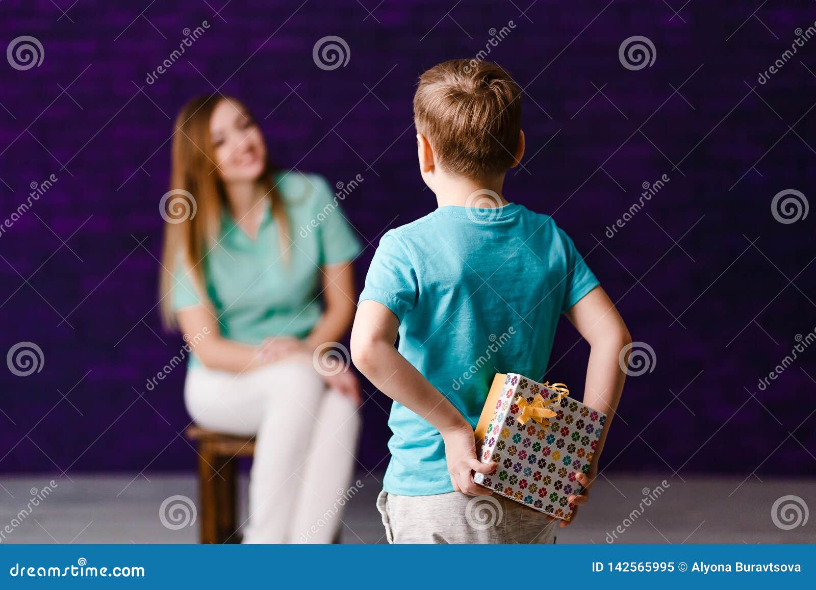Sju-år-gammal pojke som rymmer en gåvaask bak hans baksida, en ung kvinna som sitter i bakgrunden dagmamma s. Sju-år-gammal pojke som rymmer en gåvaask bak hans baksida, unga kvinnor som sitter i bakgrunden Moders dag som ger gåvor