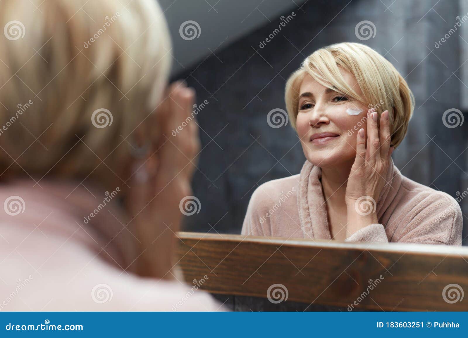 skincare routine. mature woman uses cosmetic cream on face skin in front of the mirror reflection.