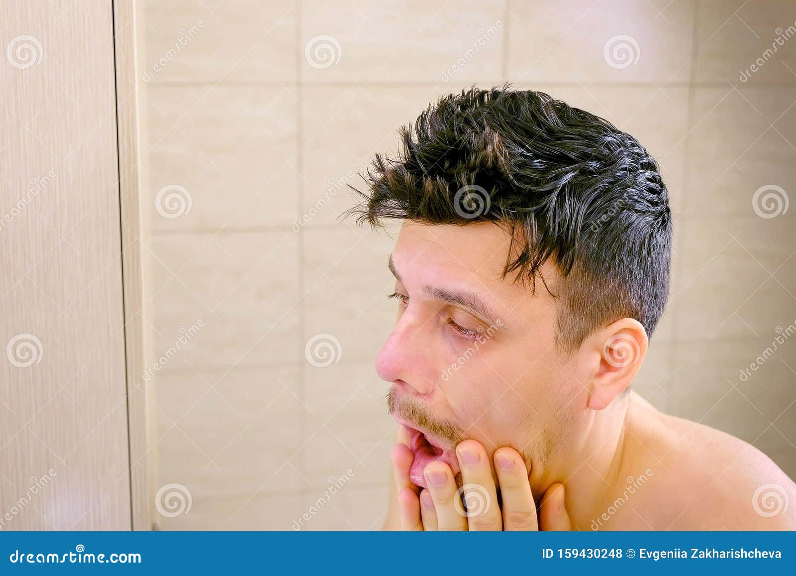 sleepy shaggy sick young man with flu looks at mirror in bathroom in morning.