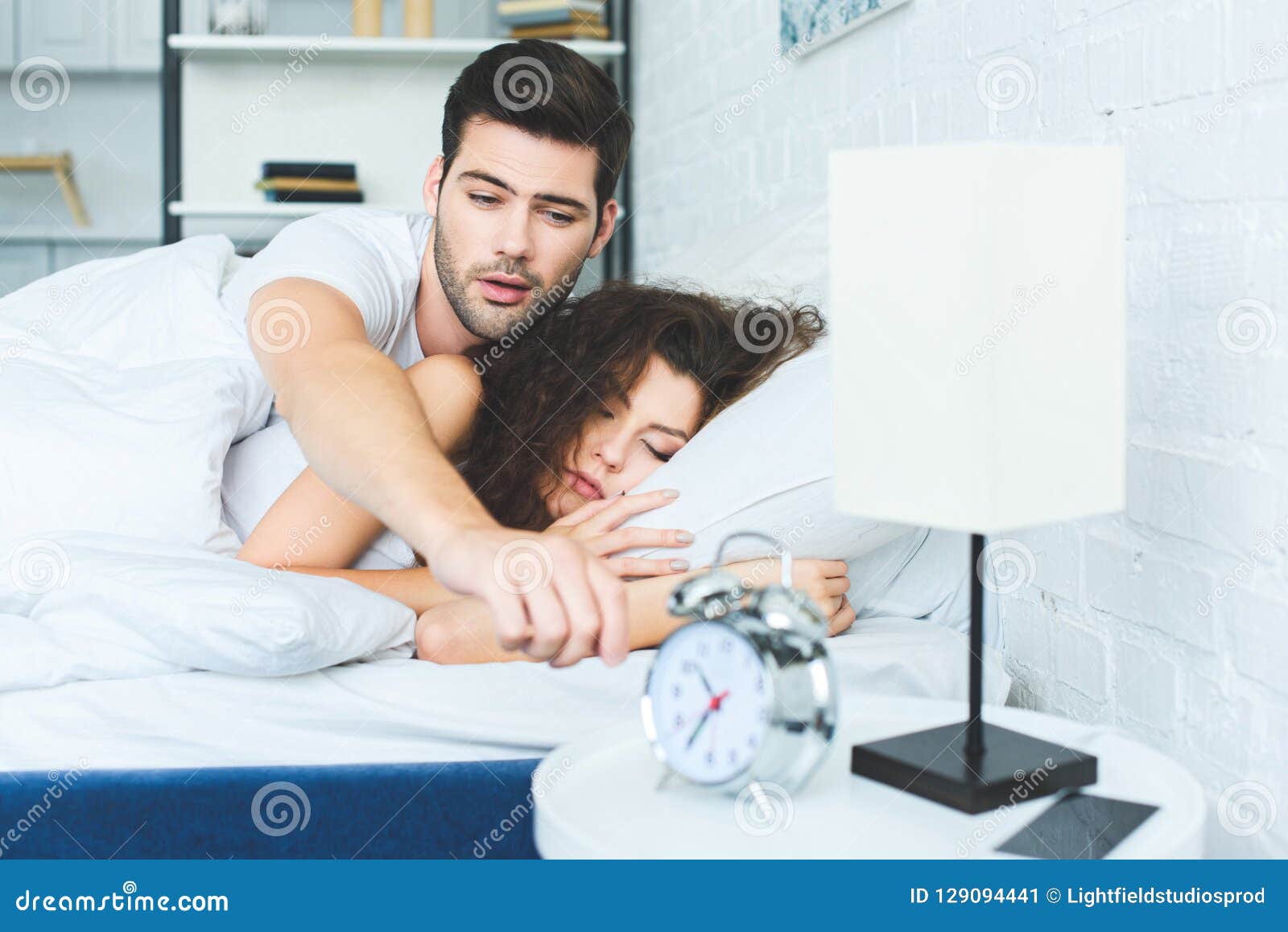 Sleepy Young Man Reaching For Alarm Clock While Sleeping With