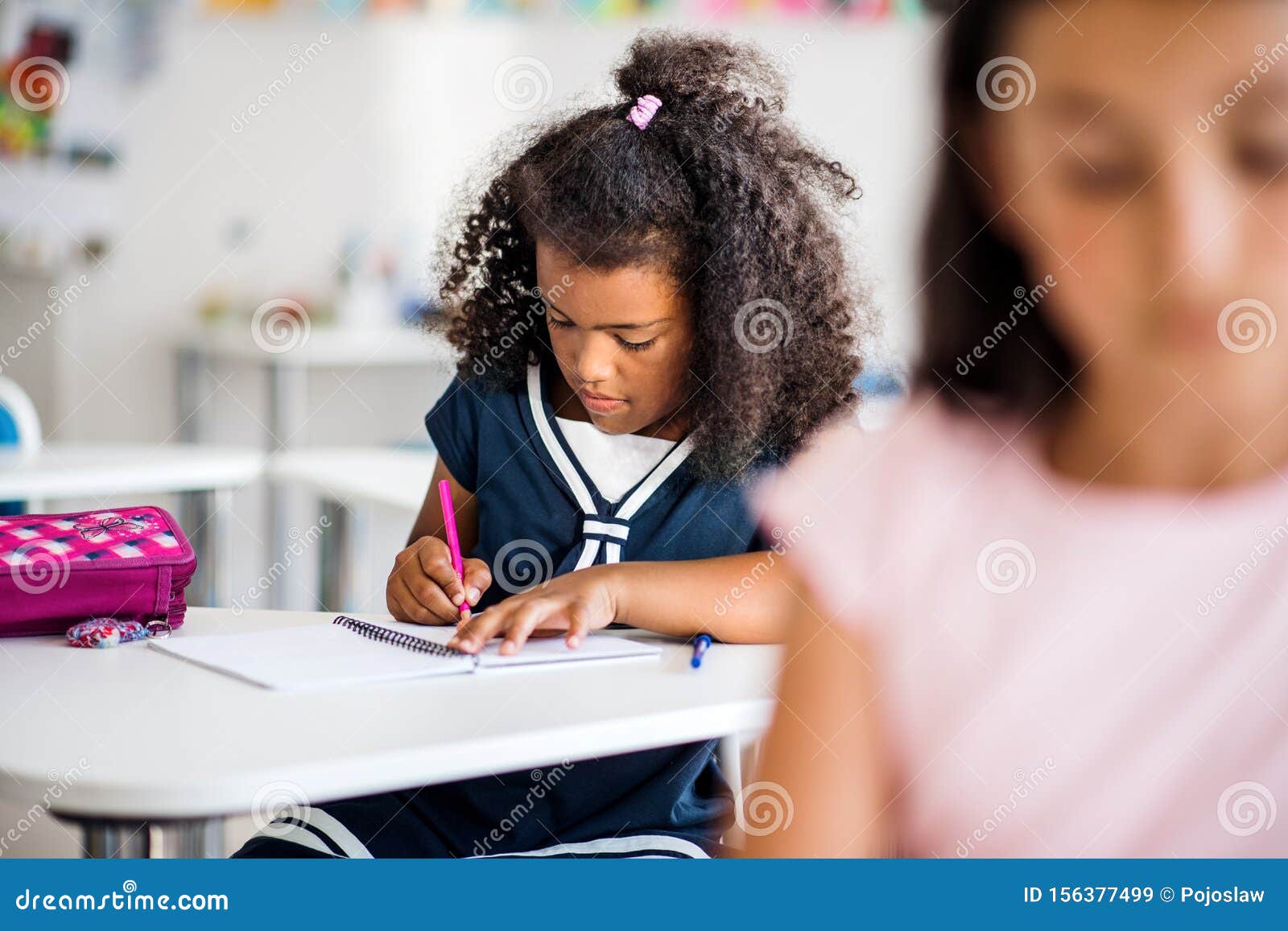 A Small School Girl Sitting At The Desk In Classroom Writing