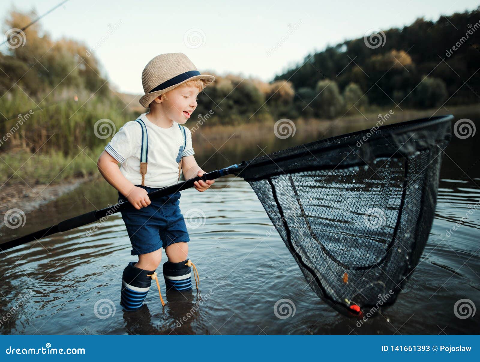 Funny happy little kid fishing on weekend. A fisherman boy stands in the  lake with a fishing rod and catches fish. Stock Photo