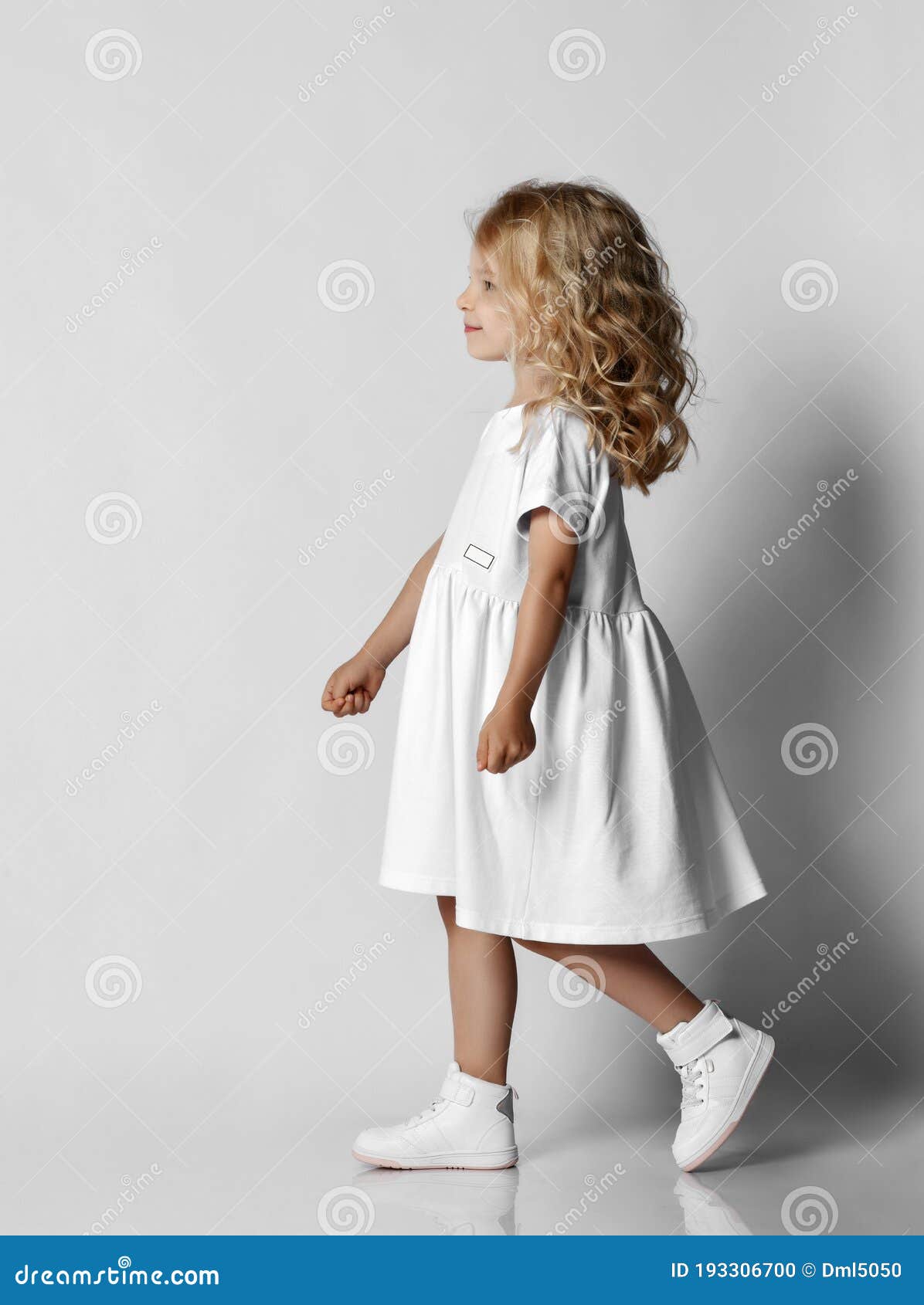 Cute Smiling Blonde Curly Kid Girl in White Casual Dress and Sneakers is  Walking, Passing Us Over Grey Background Stock Photo - Image of blonde,  girl: 193306700