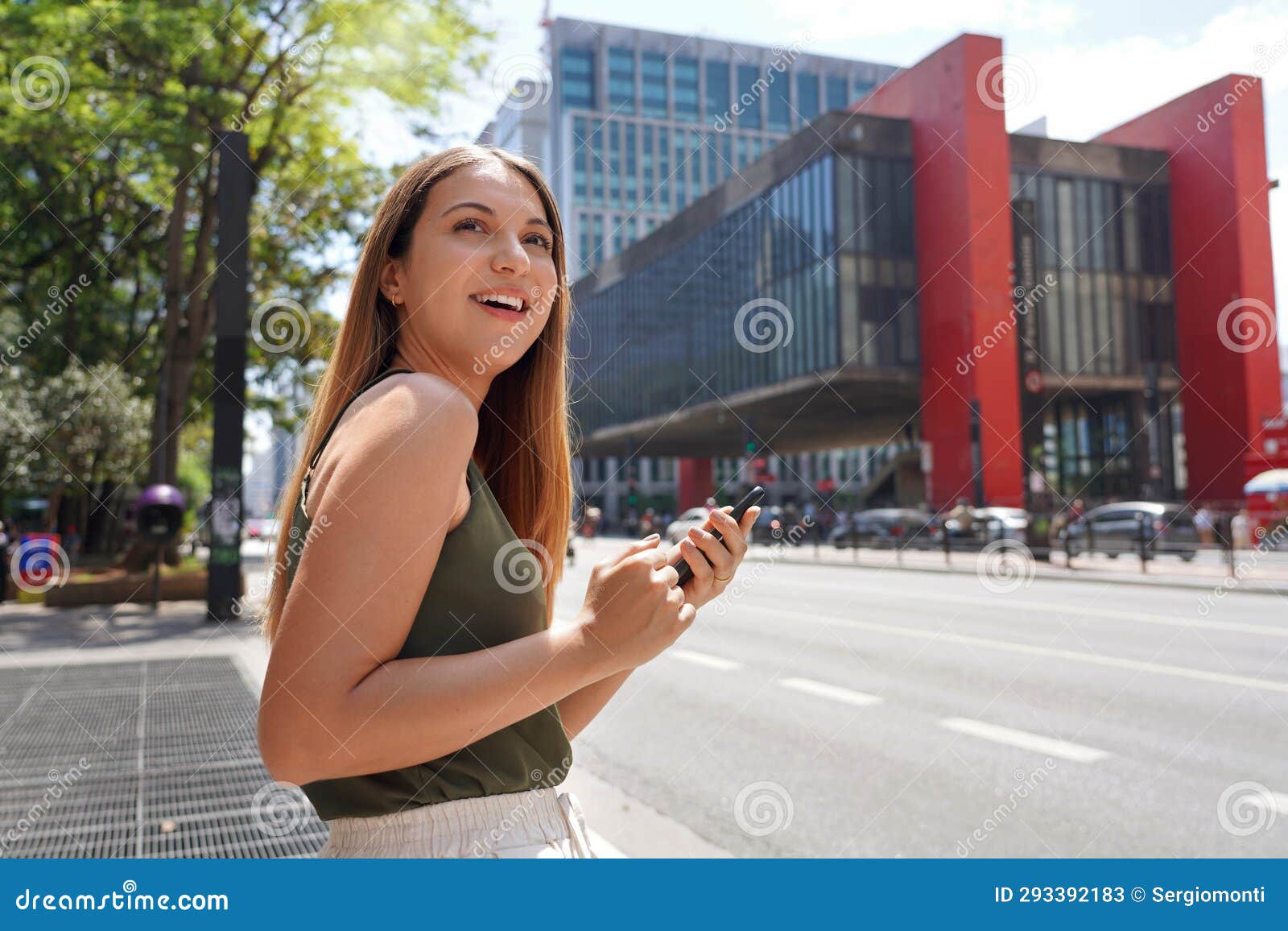 smiling brazilian woman hail a vehicle using mobile app waiting for taxi or uber on paulista avenue, sao paulo, brazil