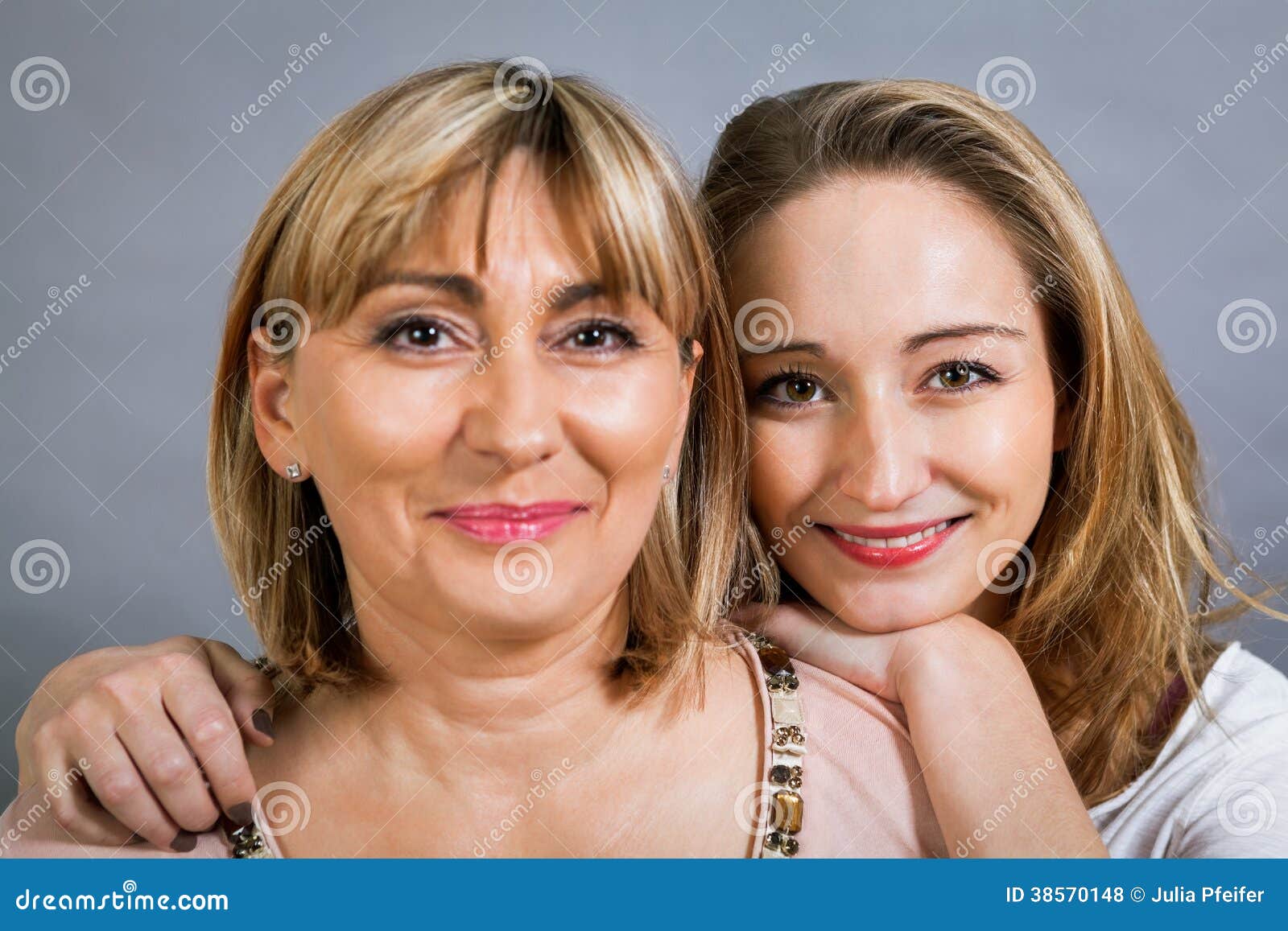 Smiling middle-aged young mother and daughter. Playful beautiful young mother and her teenage daughter posing together with the young girl peeking out to the side with a happy grin, isolated on a grey studio background