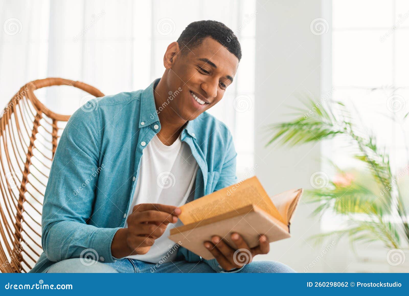 smiling smart millennial african american guy reading book, sit on chair, enjoy comfort and free time