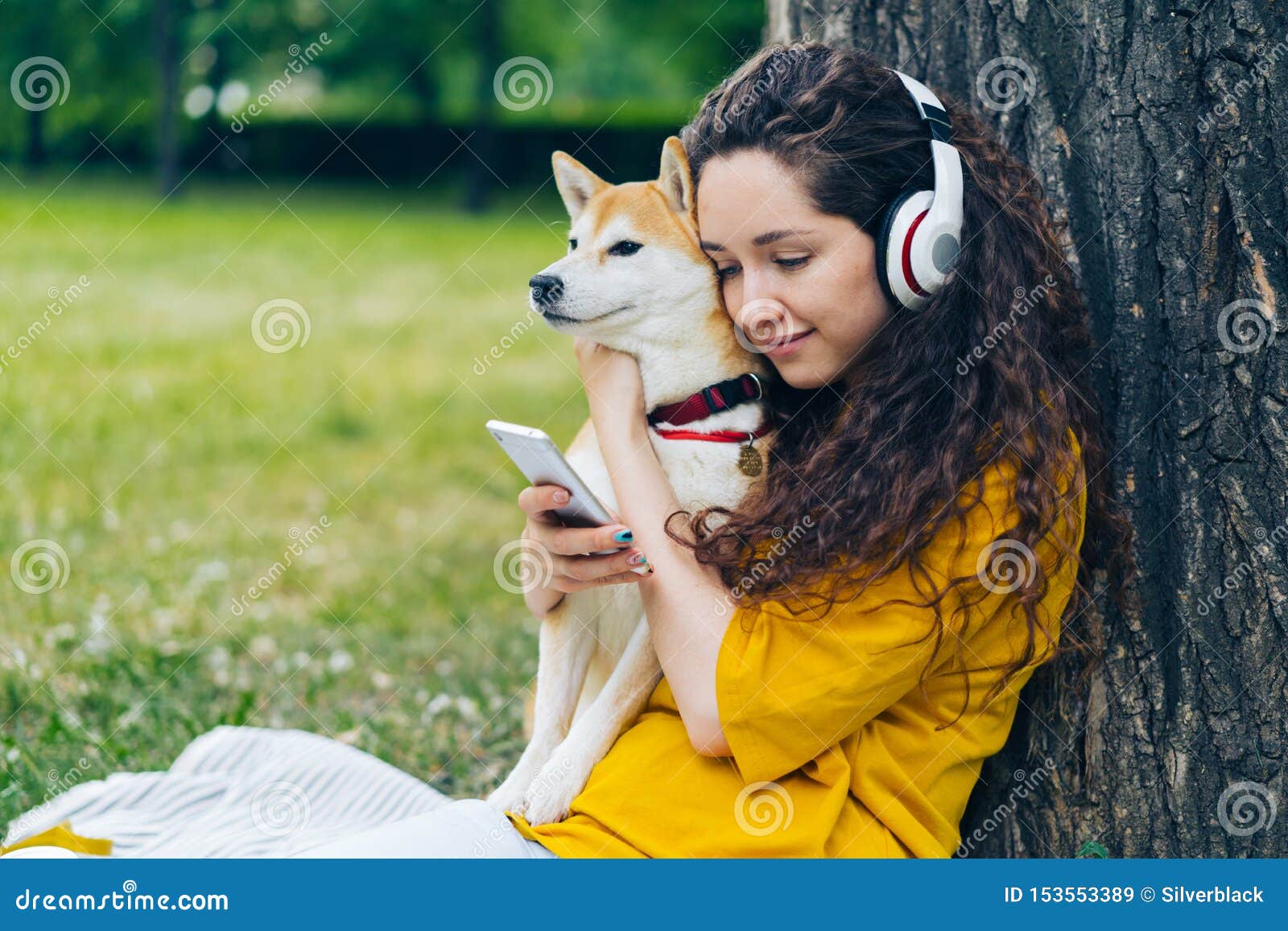 Smiling student enjoying music in headphones using smartphone in park with dog. Smiling student attractive girl is enjoying pop music in headphones using smartphone sitting on lawn in park with beautiful shiba inu dog. People and pets concept.