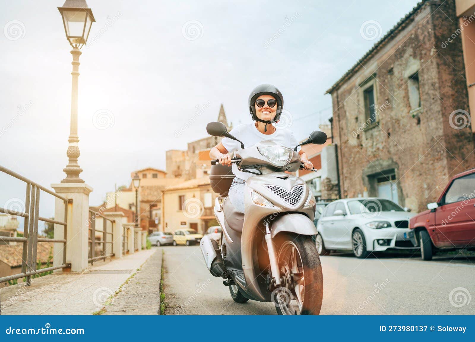 smiling woman in helmet and sunglasses fast riding the moto scooter on the sicilian old town streets in the forza d'