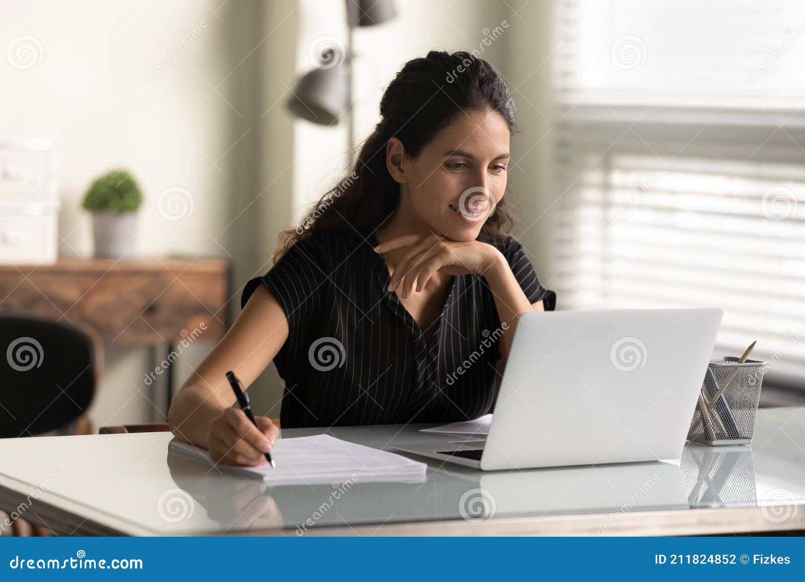 smiling woman looking at laptop screen, taking notes, studying online