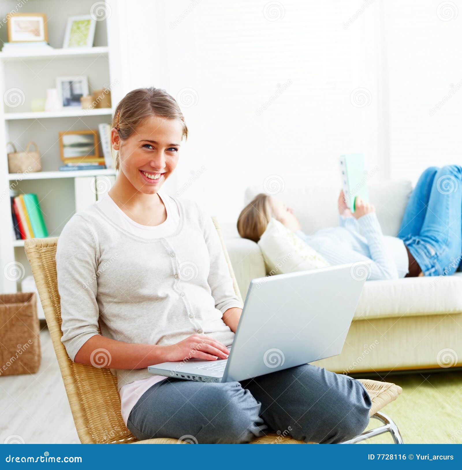 Smiling woman using laptop at home in collage dorm. Smiling woman using laptop while her friend lying on sofa reading a book