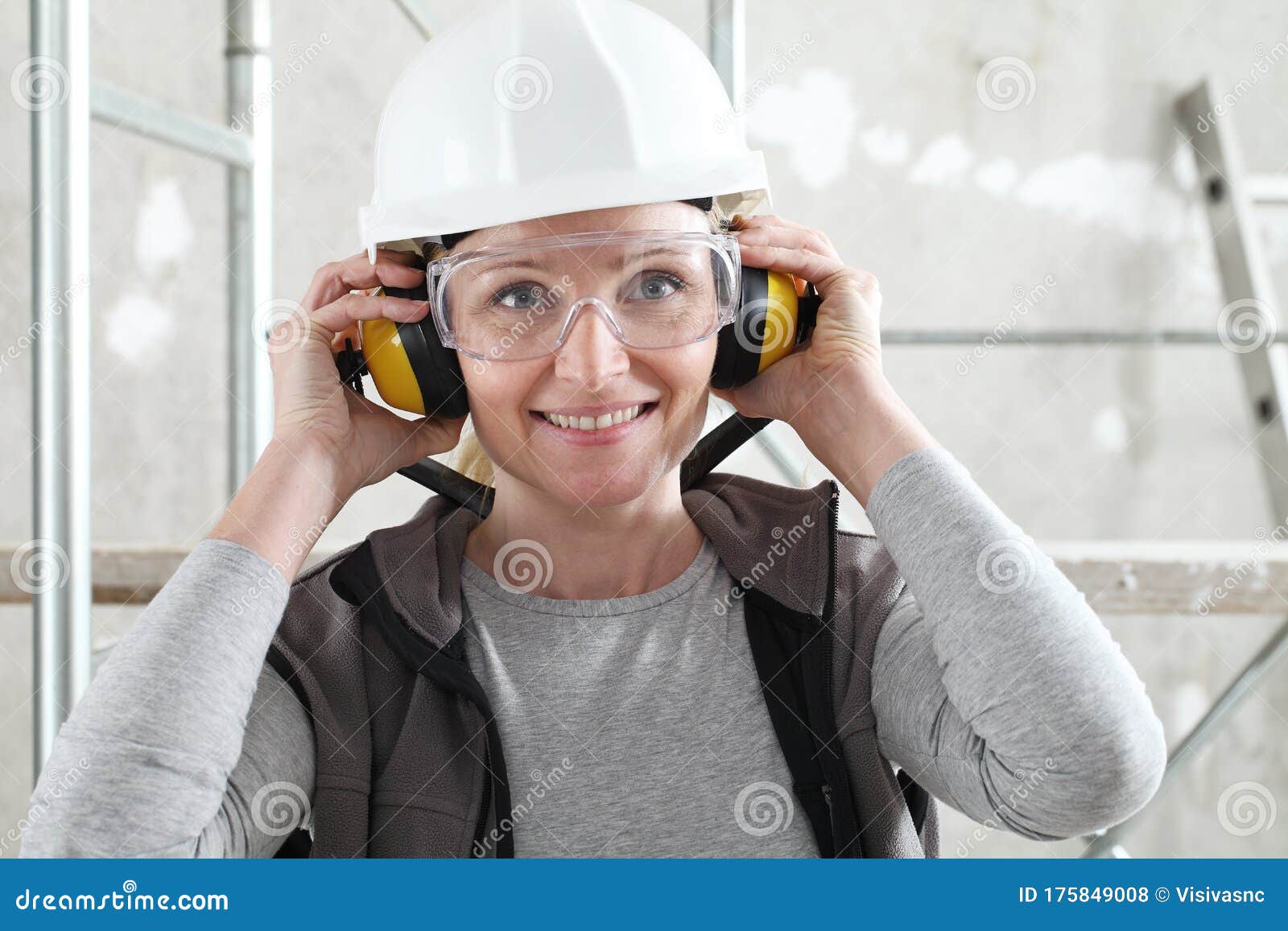 smiling woman worker portrait wearing helmet, safety glasses and hearing protection headphones, scaffolding interior construction