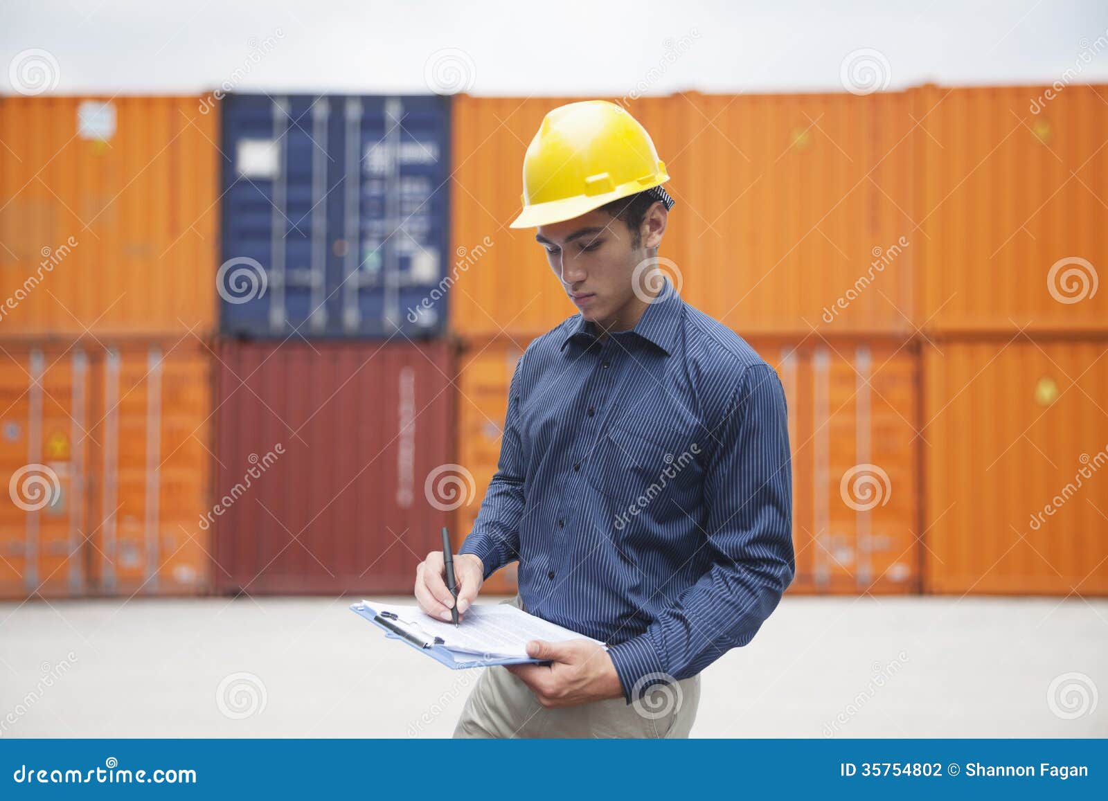Smiling Young Engineer in Protective Work Wear in a Shipping Yard Examining Cargo and Writing on
