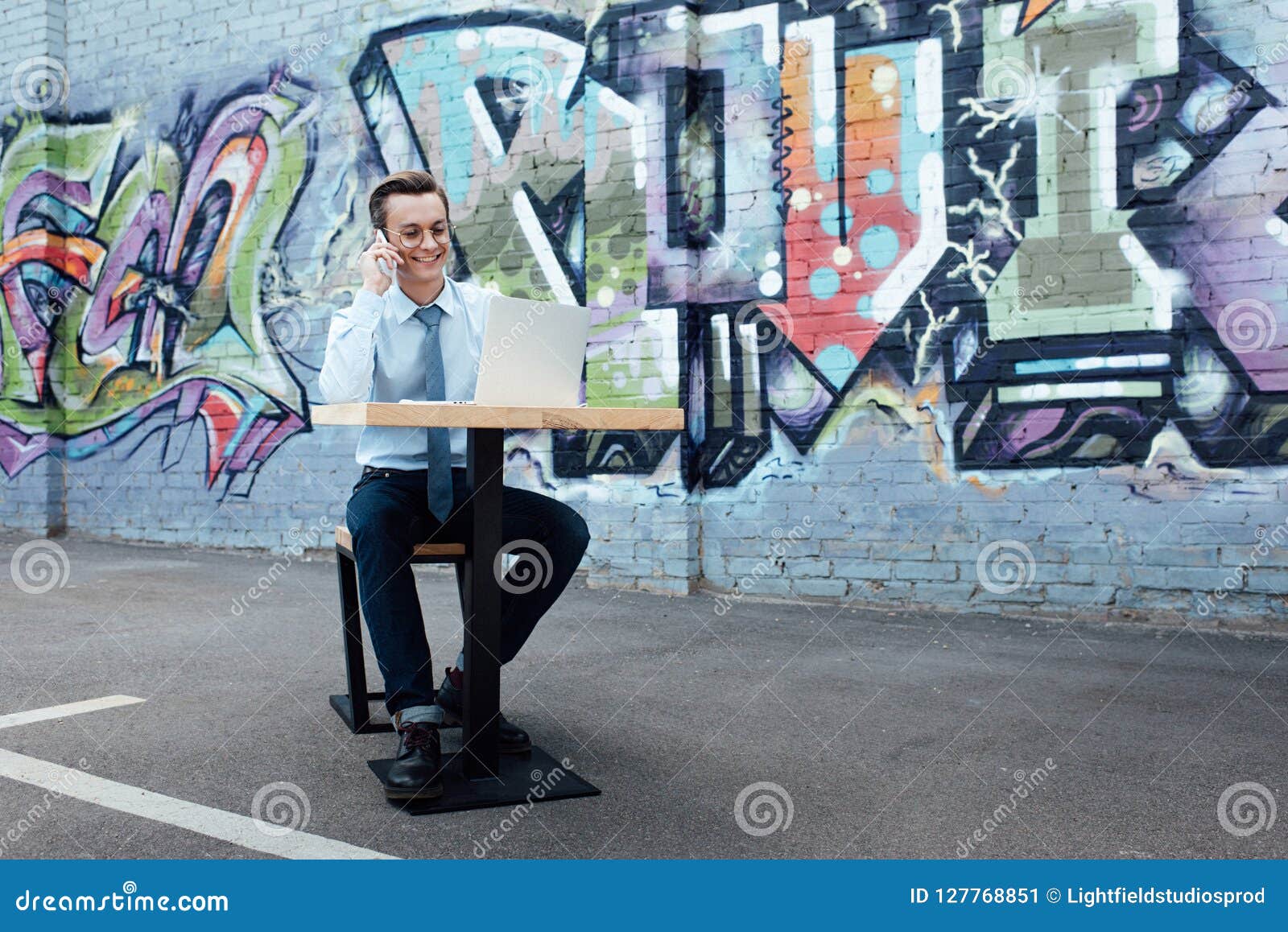 smiling young freelancer in eyeglasses talking by smartphone and using laptop while sitting