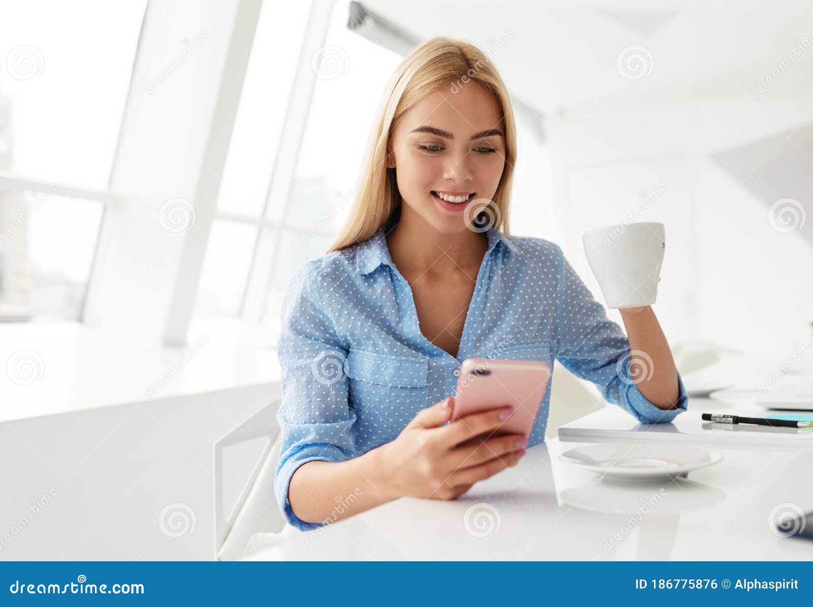 smiling young woman with cellphone in hand while drinking coffee in a white room