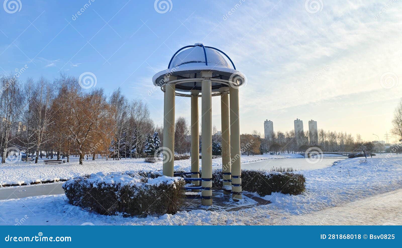 in a snow-covered city park, a rotunda of concrete columns with a dome-d roof stands next to a pedestrian path and an ice-cov