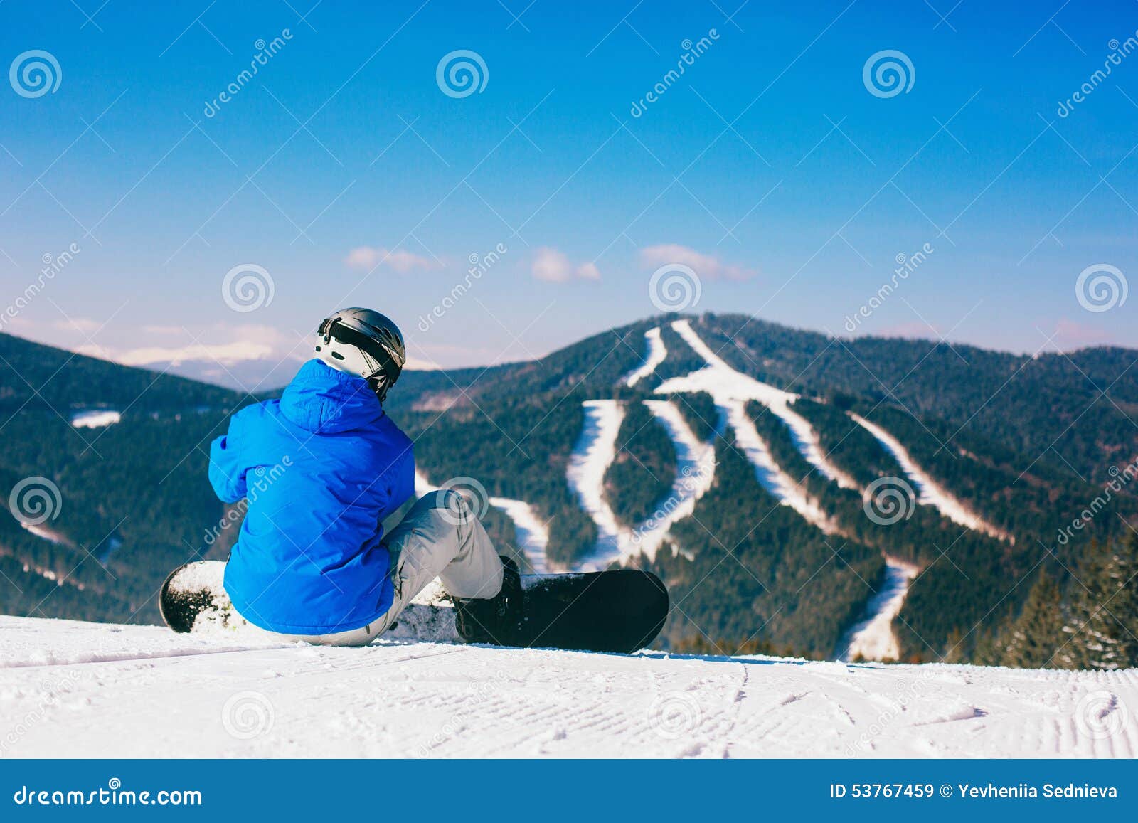Snowboarder que senta-se na neve contra montanhas e floresta do abeto