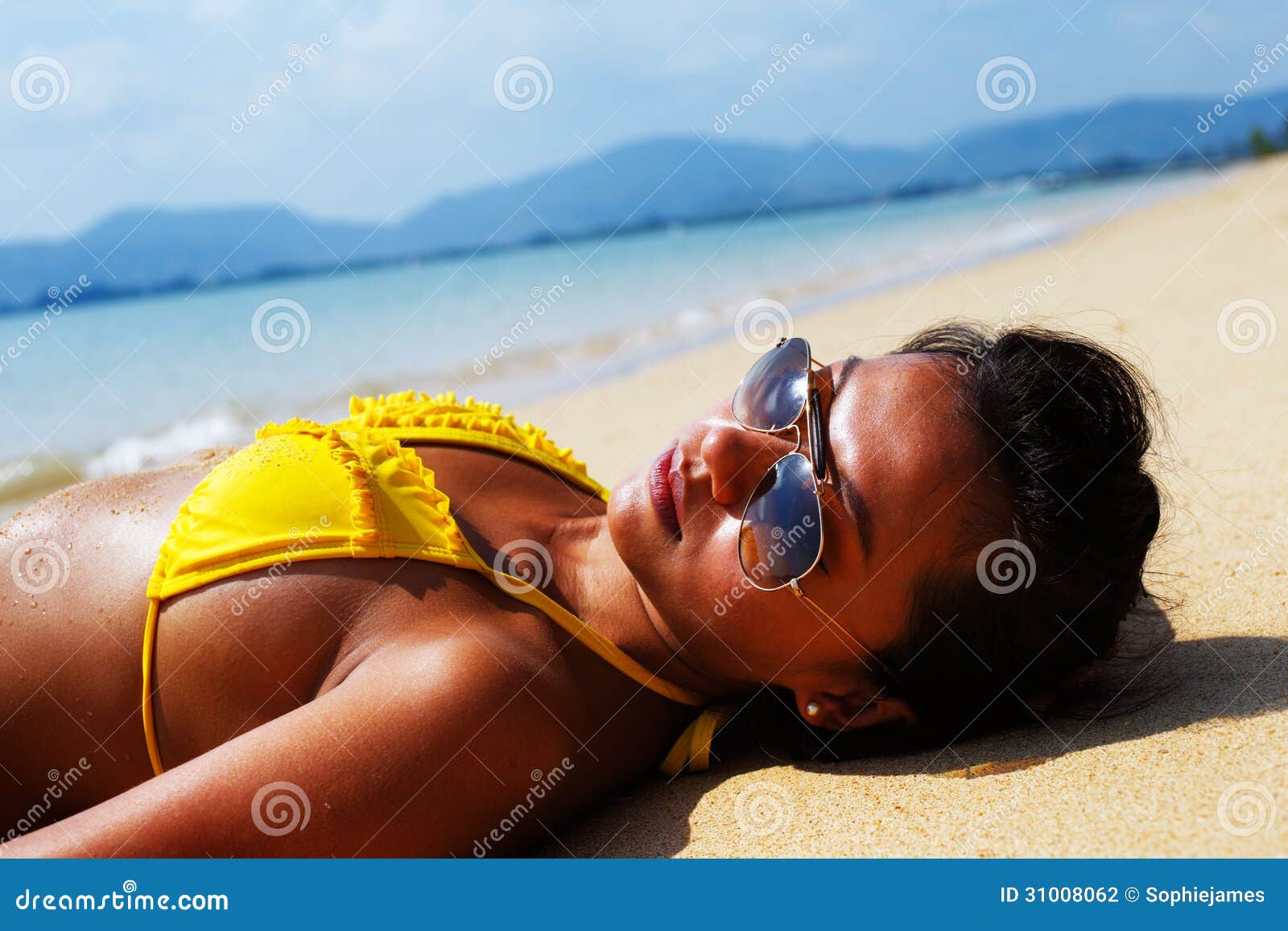 Solbadning för ung kvinna på en sandig strand av Thailand. Ung kvinna i en gul baddräkt och solglasögon som lägger ner på en sandig strand av Thailand på en solig sommardag