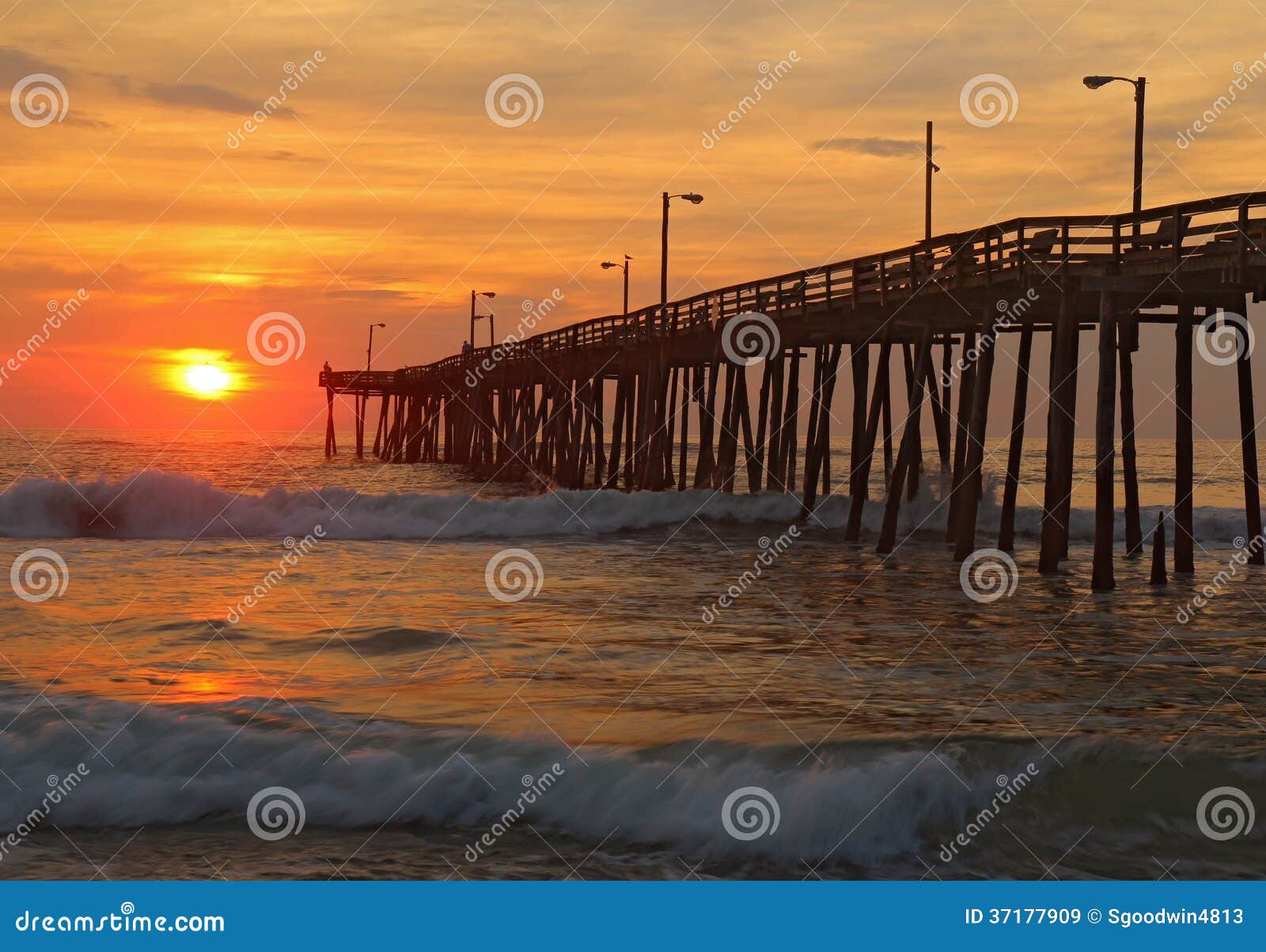 Sonnenaufgang durch einen Fischenpier im North Carolina. Das aufgehende Sonne späht durch Wolken und wird in den Wellen durch den Nagshauptfischenpier auf den äußeren Banken des North Carolina reflektiert