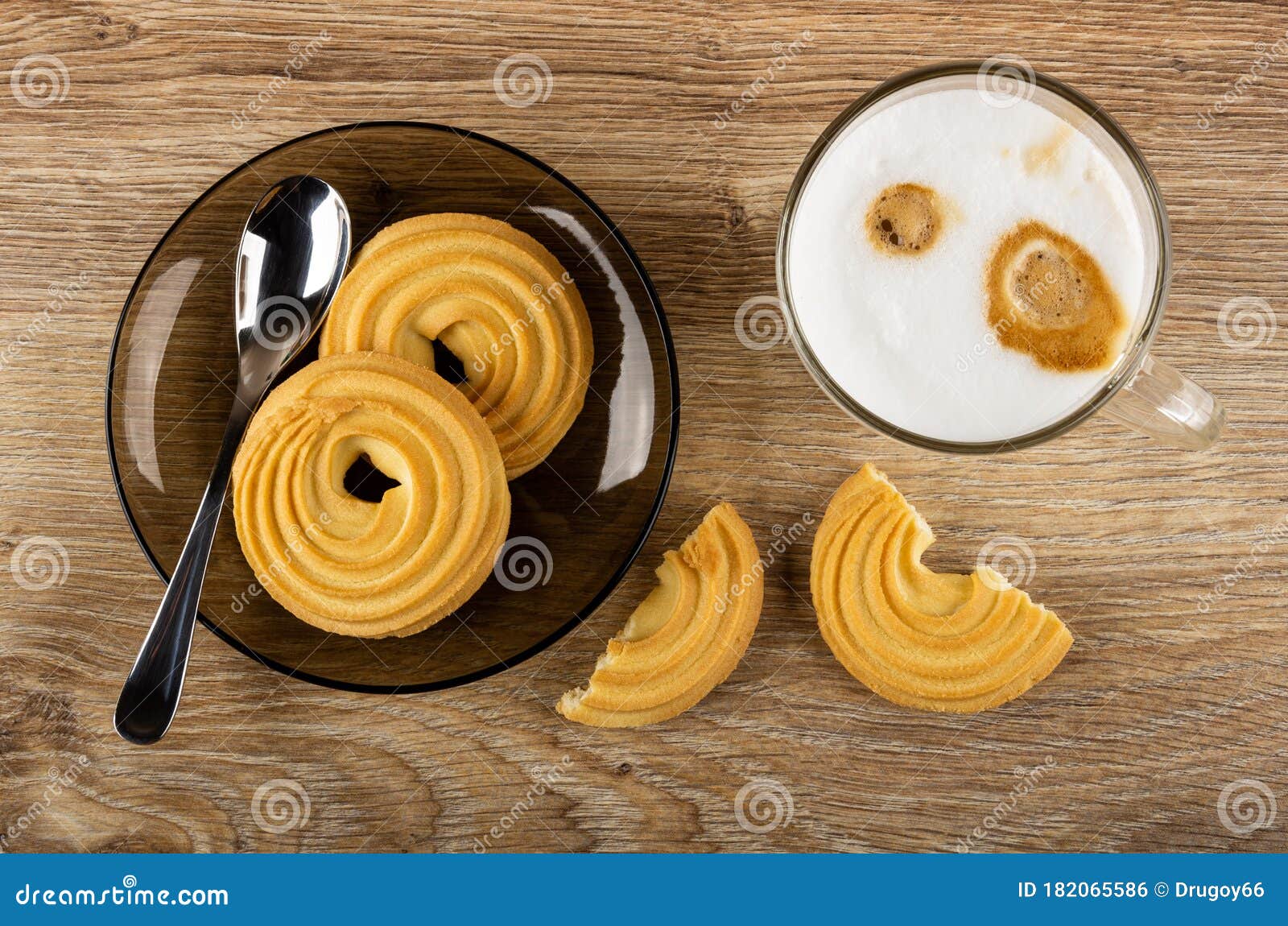 spoon, cookies in brown saucer, transparent cup with latte-macchiato, pieces of cookie on table. top view