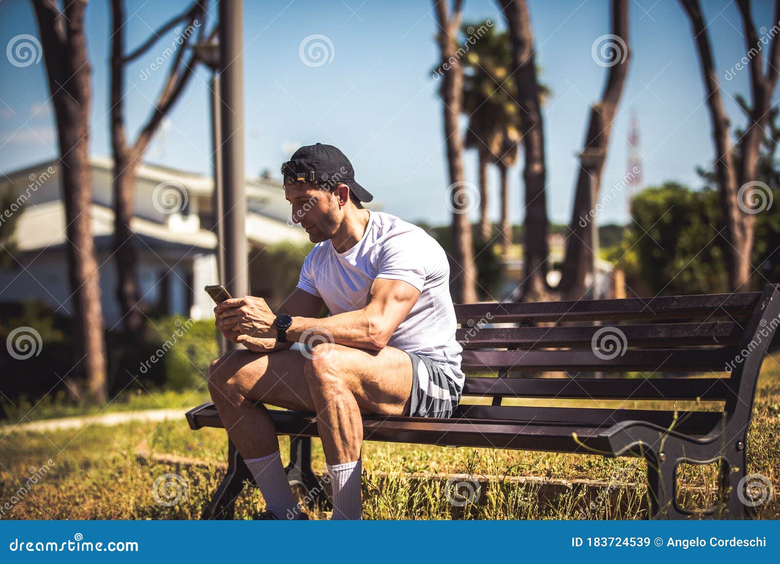 sporty man personal trainer sitting looking at his phone on a bench in an outdoor park. muscle body and well defined. sporty