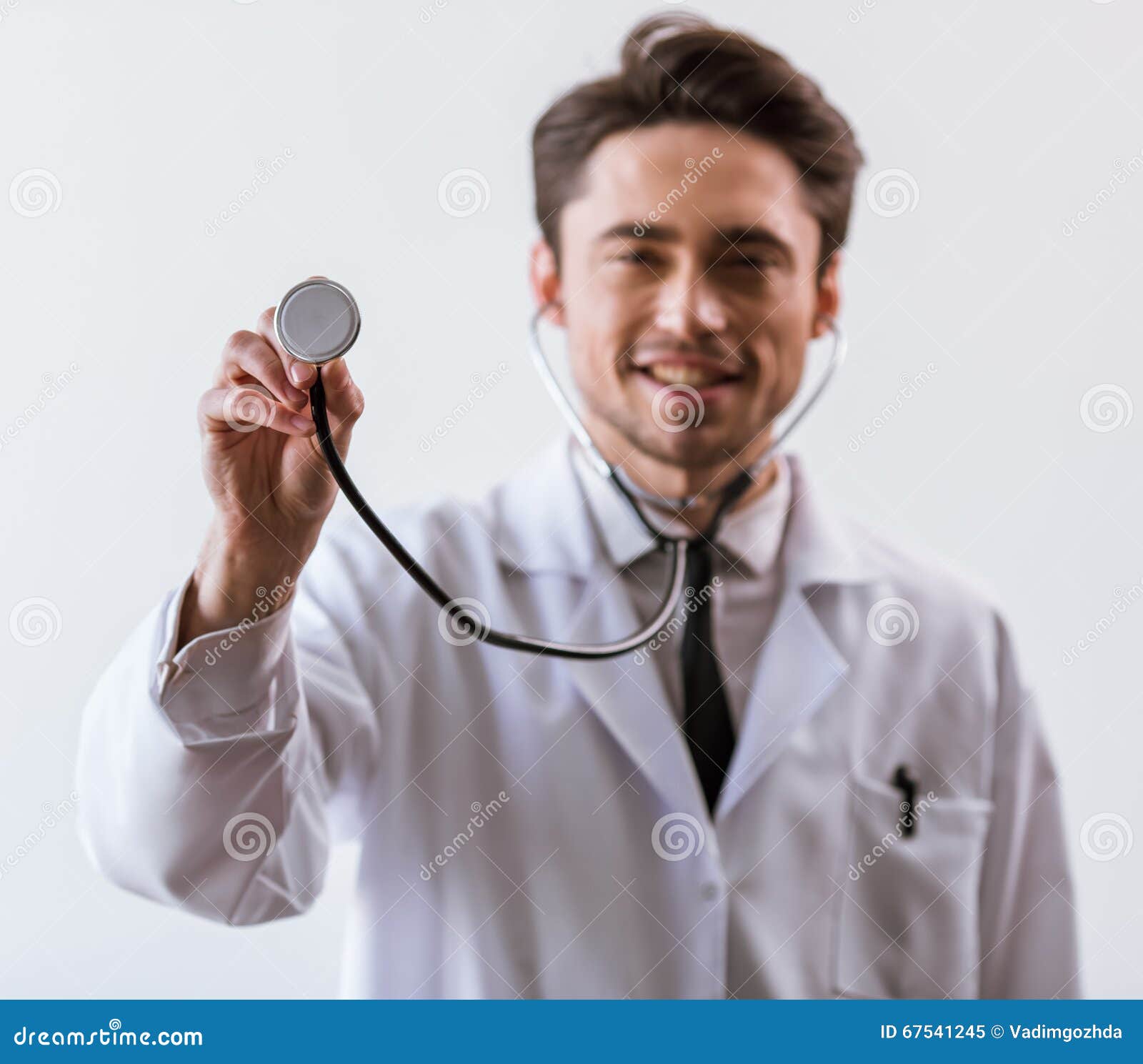 Stattlicher junger Doktor. Hübscher junger Doktor im weißen Kleid und im Stethoskop lächelnd, Kamera betrachtend und Stethoskop zeigend, konzentrieren sich an Hand, auf weißen Hintergrund