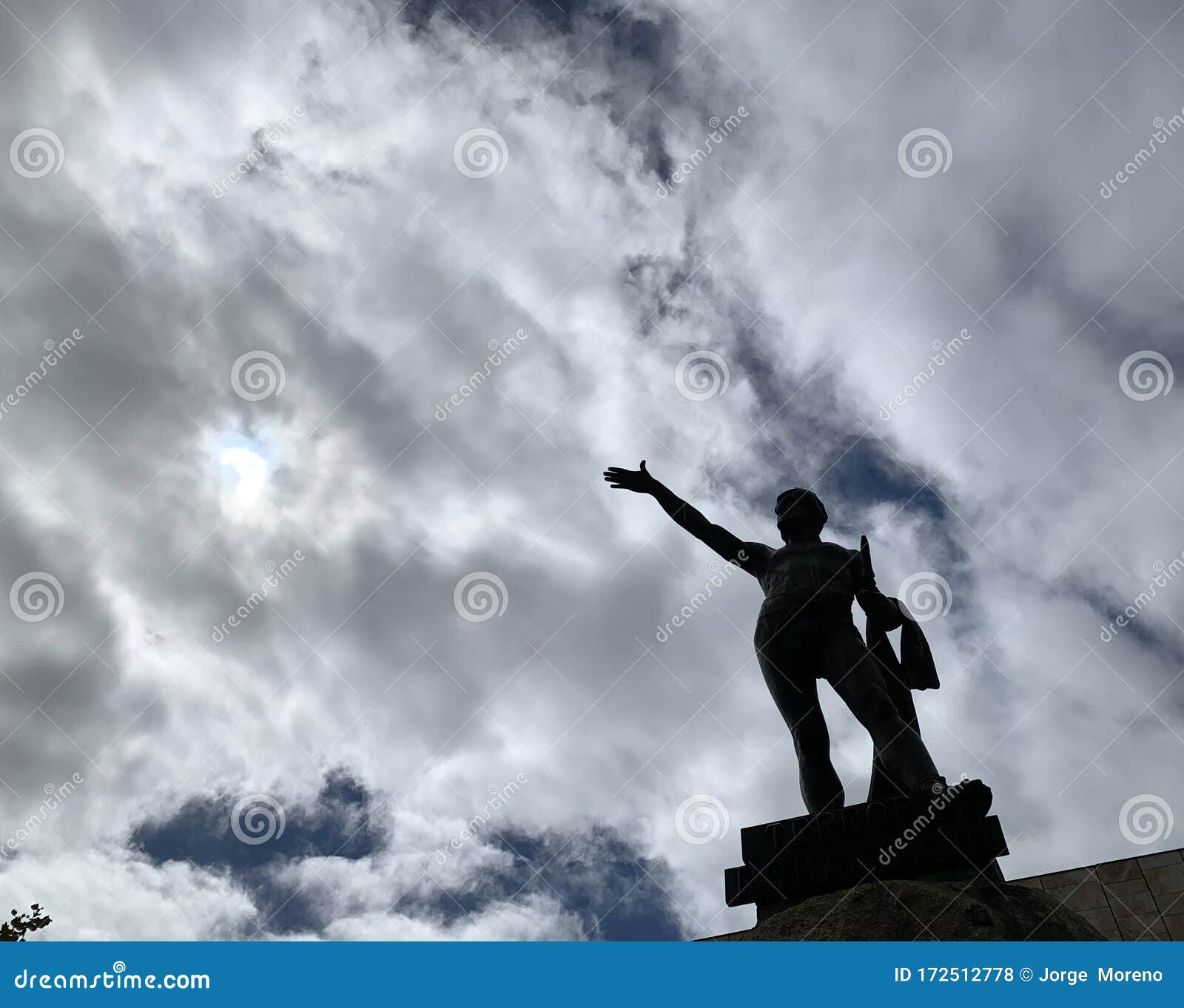 a statue in the square of a town in spain on a cloudy day in the light of noon