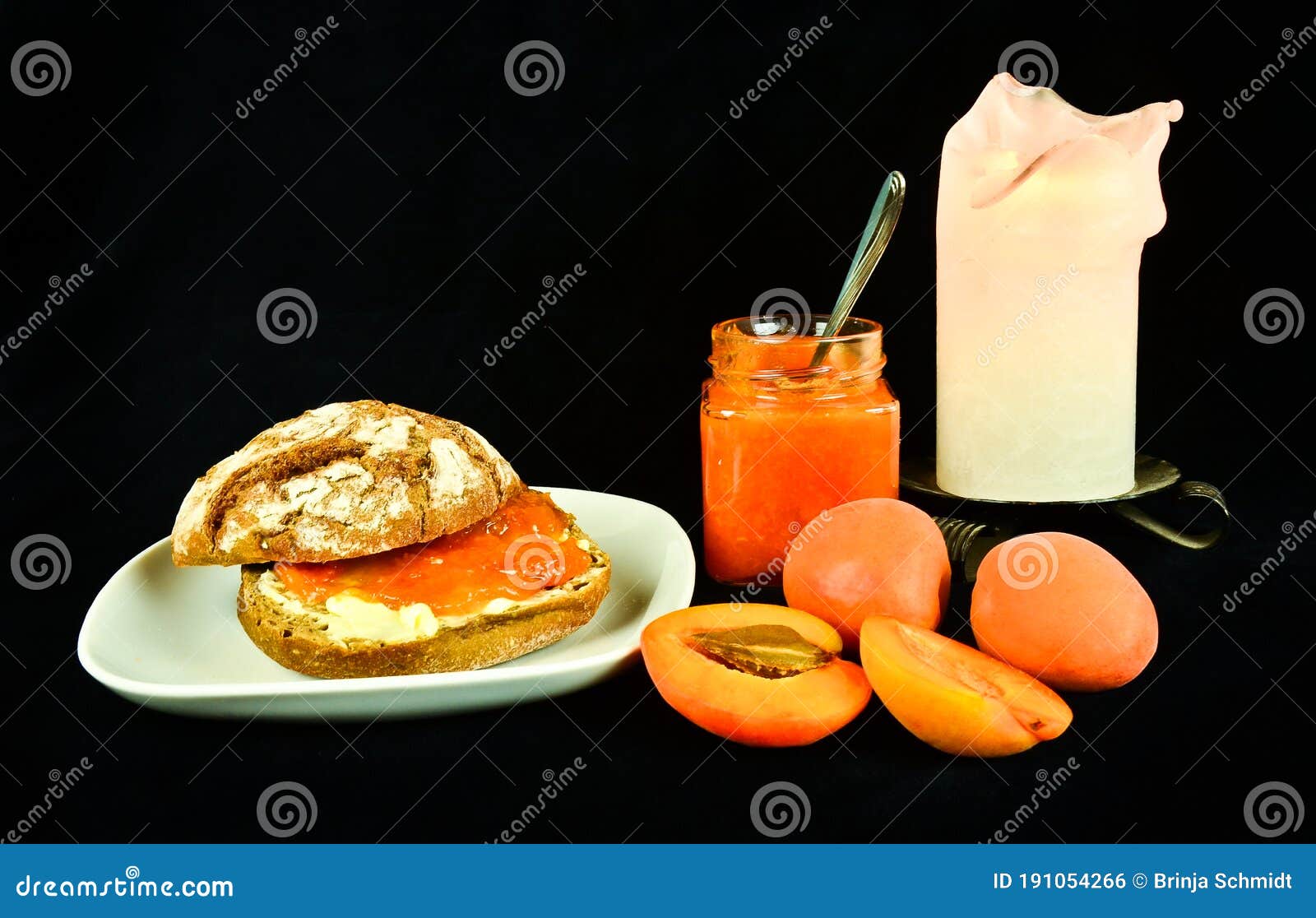 a still life with apricots, apricot jam and a bread roll with butter and jam ready to eat in front of black background