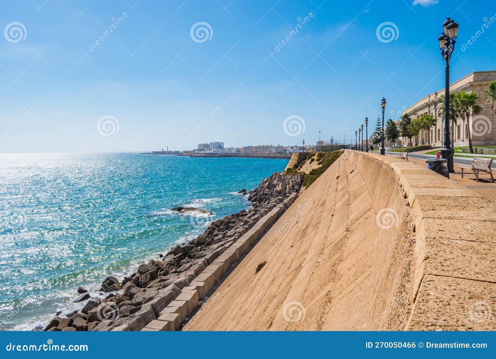 stone wall and breakwater with blue sea and coastline of cÃ¡diz with sunlight on horizon, spain