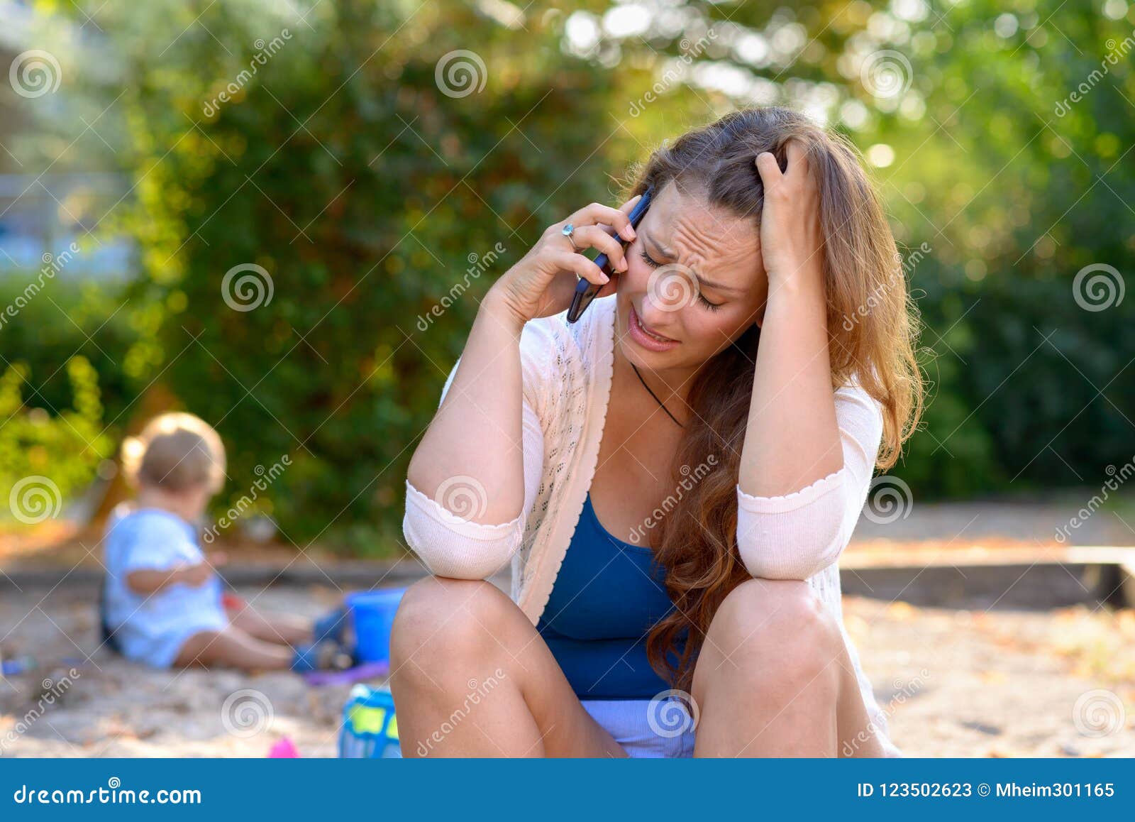 Stressed comprimiu a mãe que fala em um móbil. Stressed comprimiu a mãe nova que fala em um telefone celular como se senta fora em um campo de jogos com seu filho do bebê que joga atrás dela