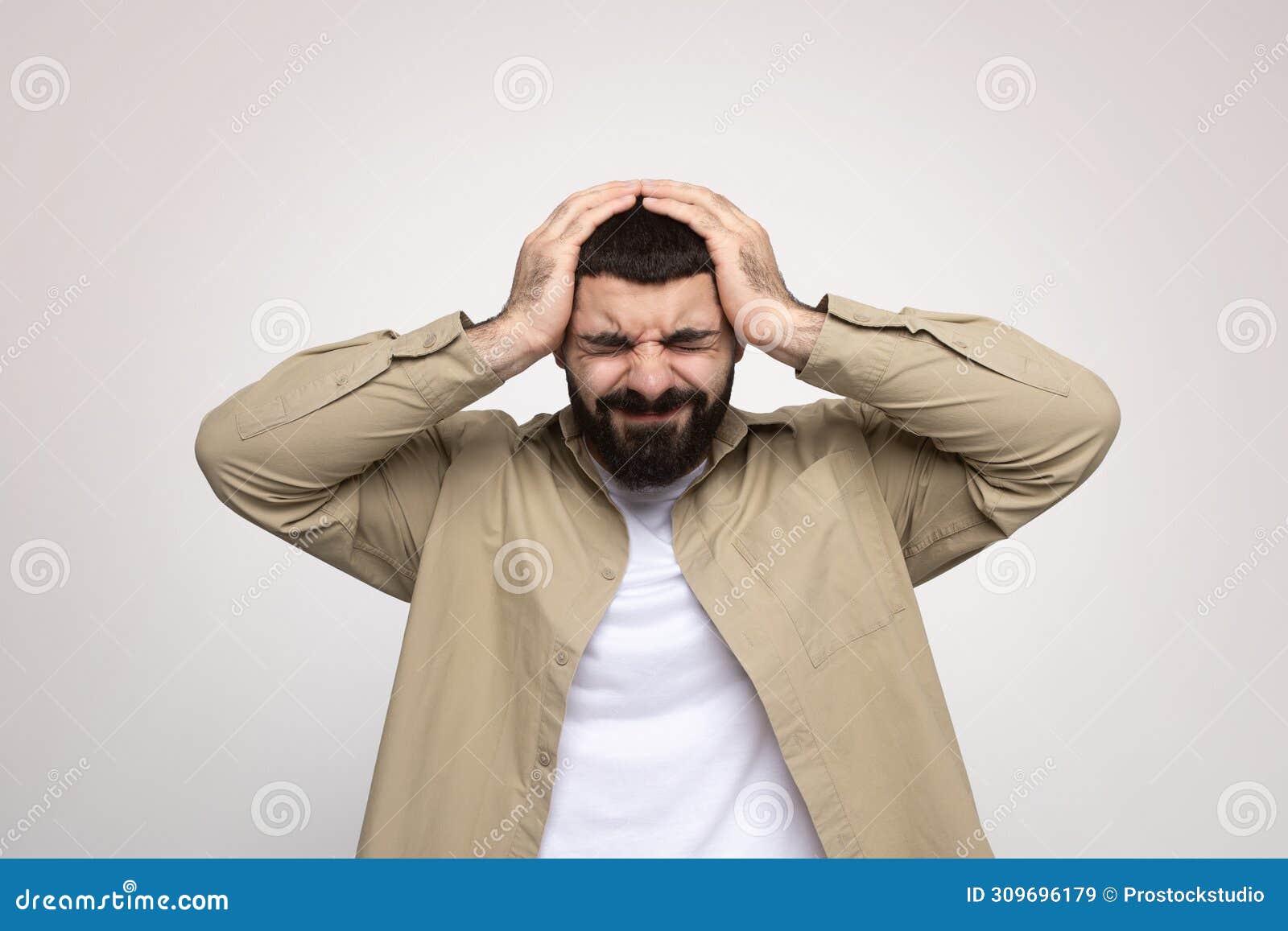 Stressed man with a beard grimacing in frustration, holding his head in his hands. Stressed millennial arab man with a beard grimacing in frustration, holding his head in his hands, wearing a beige jacket over a white t-shirt, against a light background