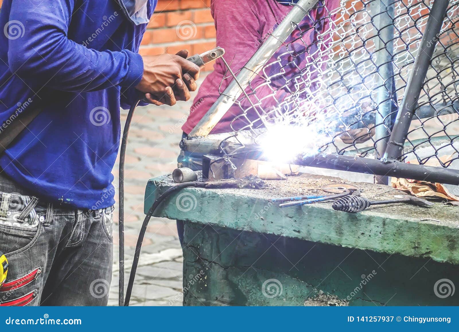 strong man is a welder in a blue t-shirt,and welders leathers, a metal product is welded with a welding machine in the outdoor