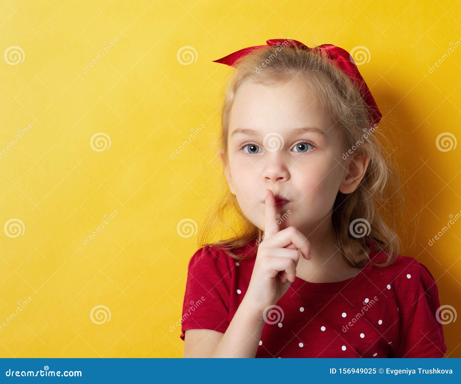 Strålningskort om en ganska ung blond flicka som visar en tyst gest och vinkar isolerad över gul bakgrund. Porträtt från en söt, blond flicka med en röd båge som visar en tyst gest, isolerad på gul bakgrund