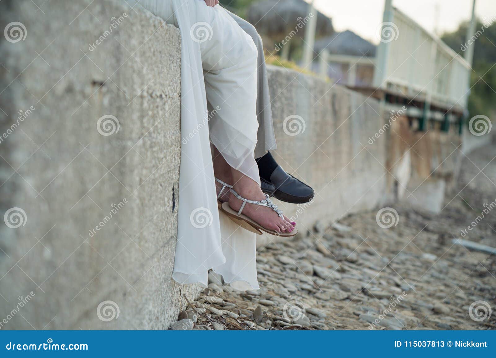 A Bride And Groom Sit On A Rock Wall By The Beach Stock