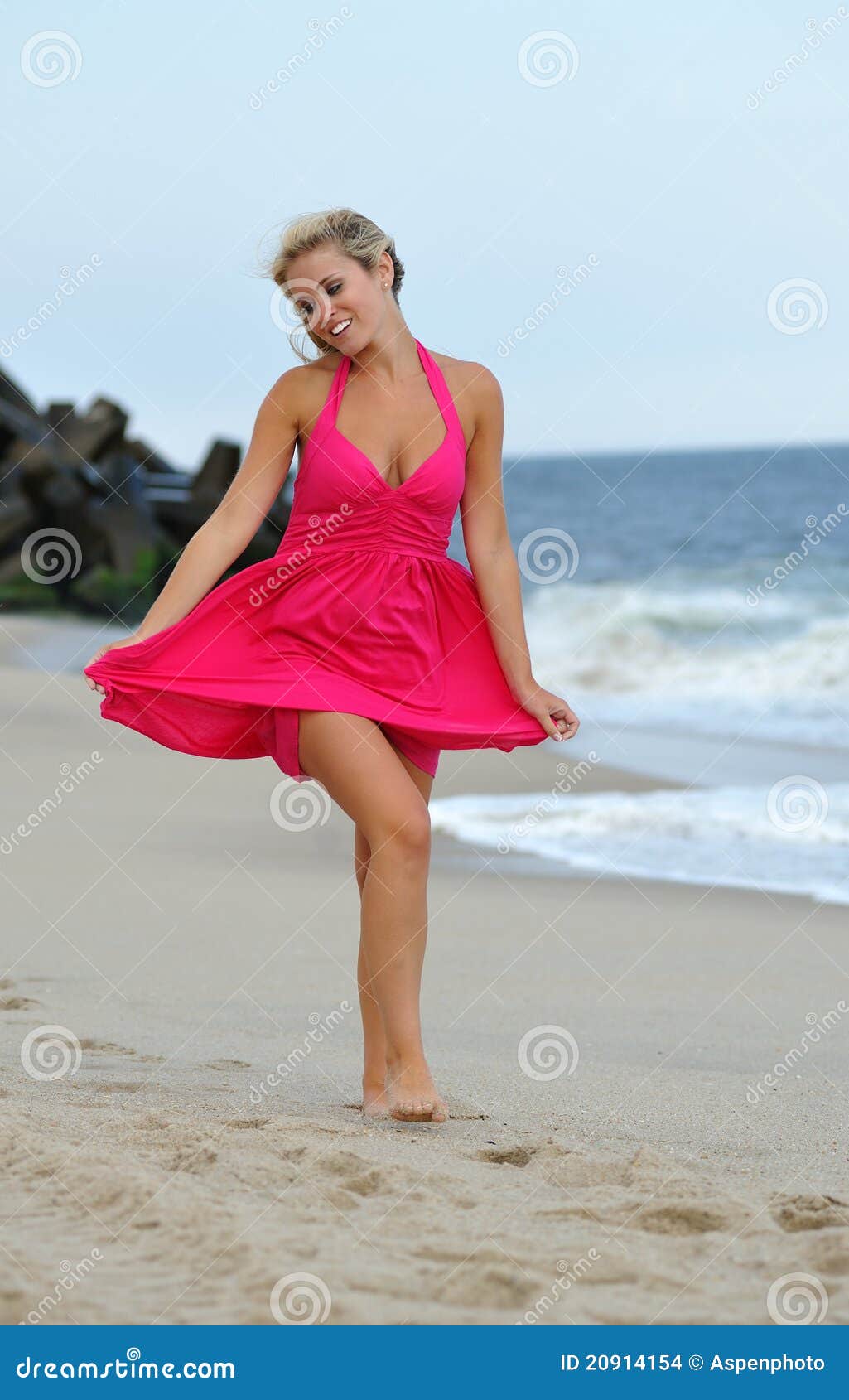 Stunning young blonde woman walking on the beach. Beautiful young blonde woman in a red sun dress walking barefoot on the beach along the waterline - stone jetty in the background. Smiling