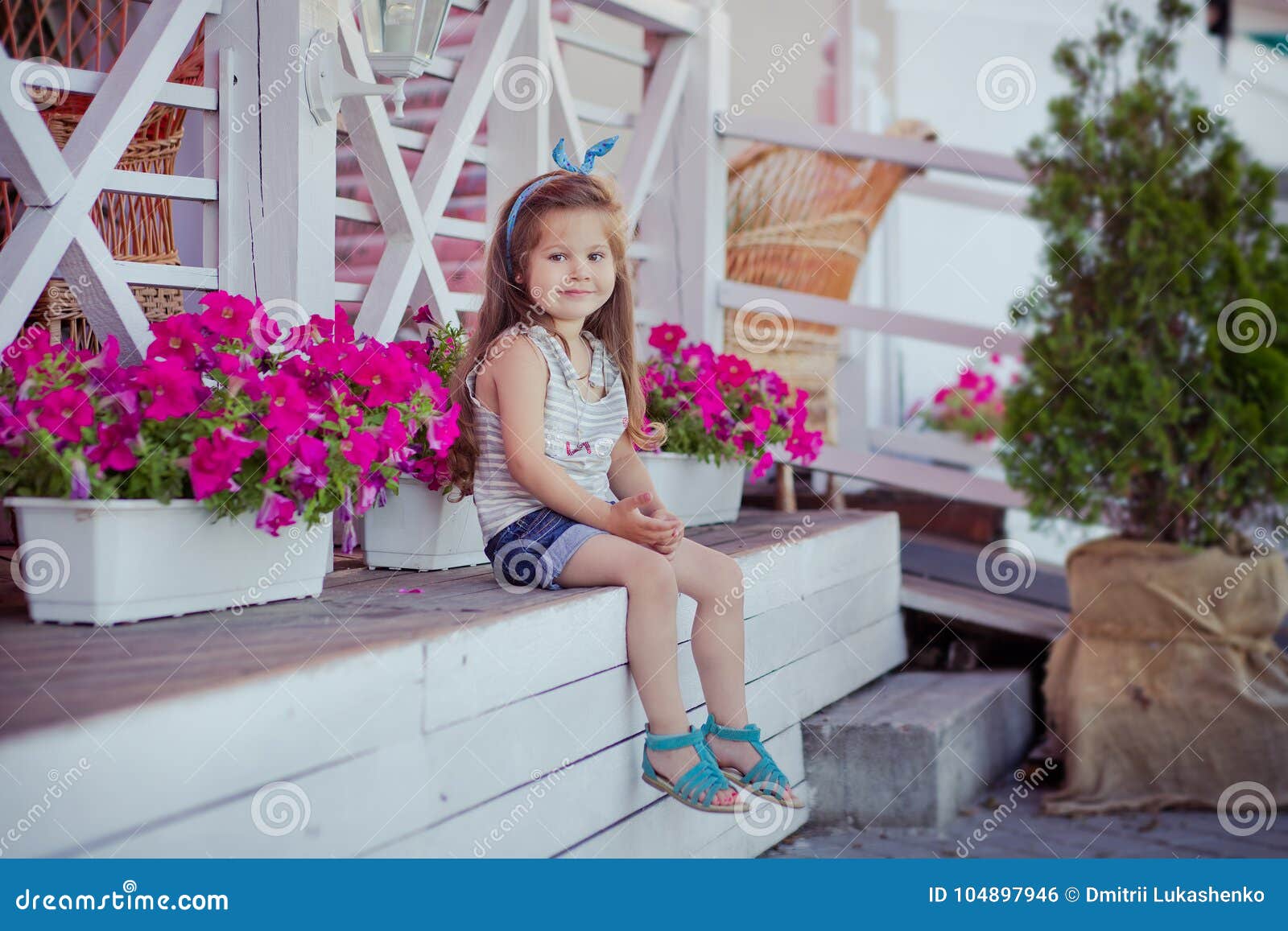 stylish beautifull cute baby girl with brunette hair posing on wooden garden full of flowers wearing tiny jeans shirts and airy sk