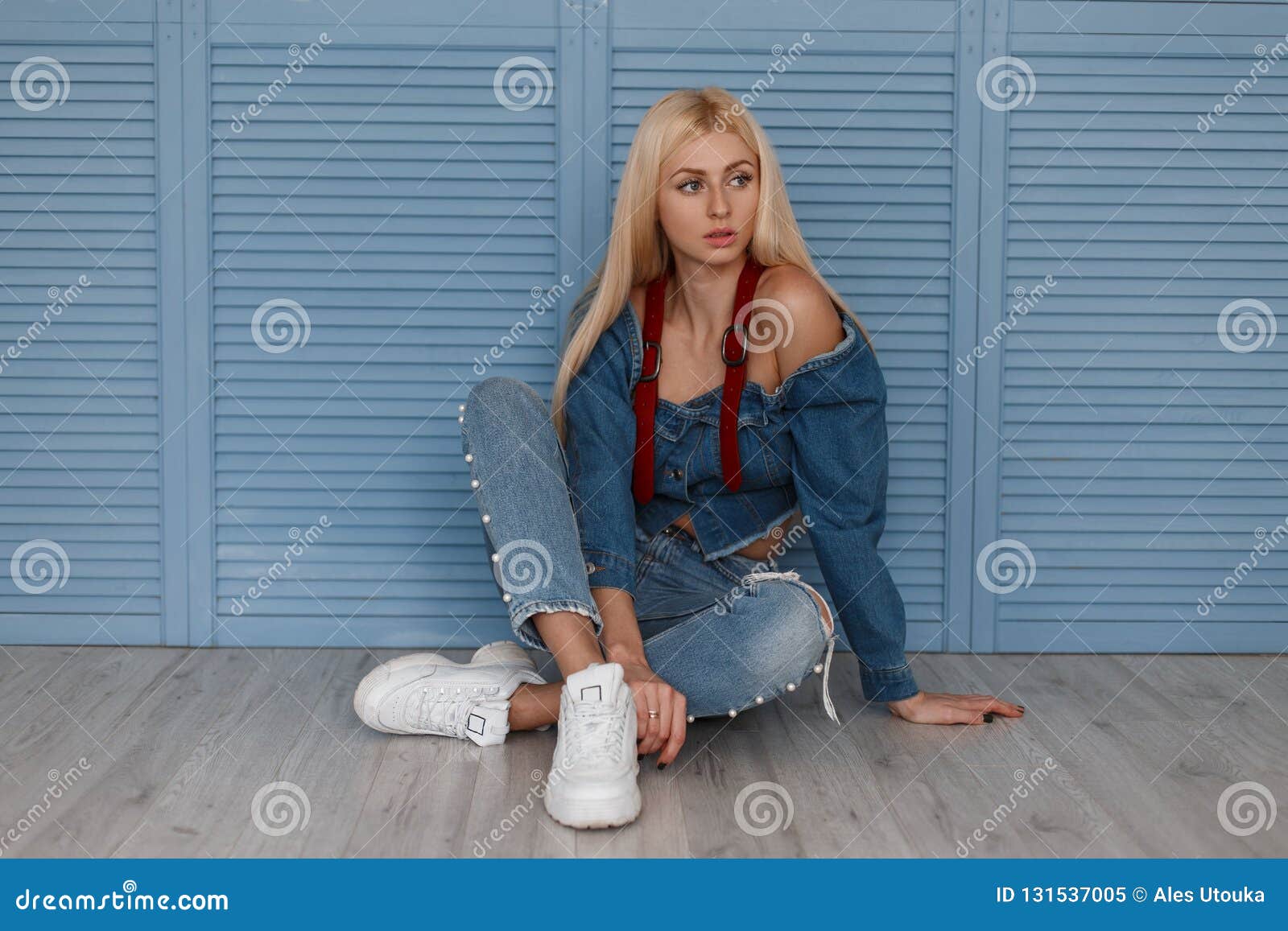 Stylish Blonde Woman In A Denim Shirt With Red Belts And Blue Jeans With White Shoes Sits Near Blue Wooden Wall Stock Image Image Of Caucasian Person