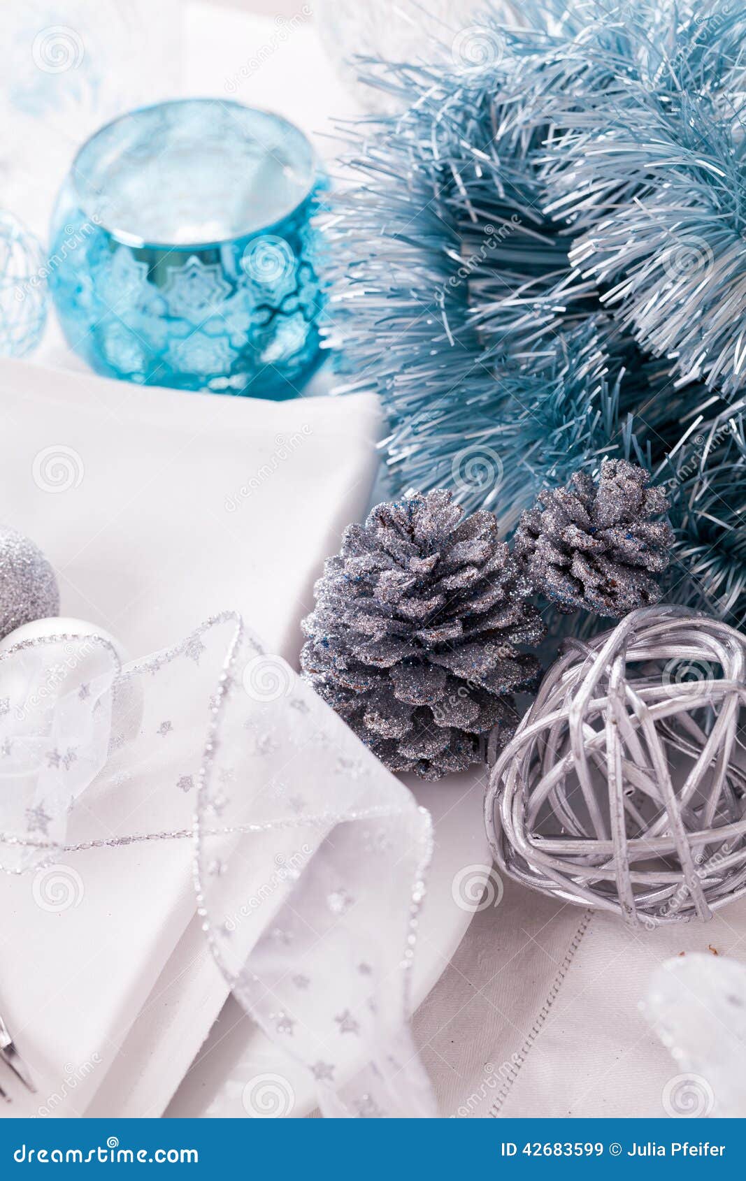 Stylish blue and silver Christmas table setting with a pretty translucent bow on white dinnerware with silver cutlery, pine cones and baubles, high angle close up view