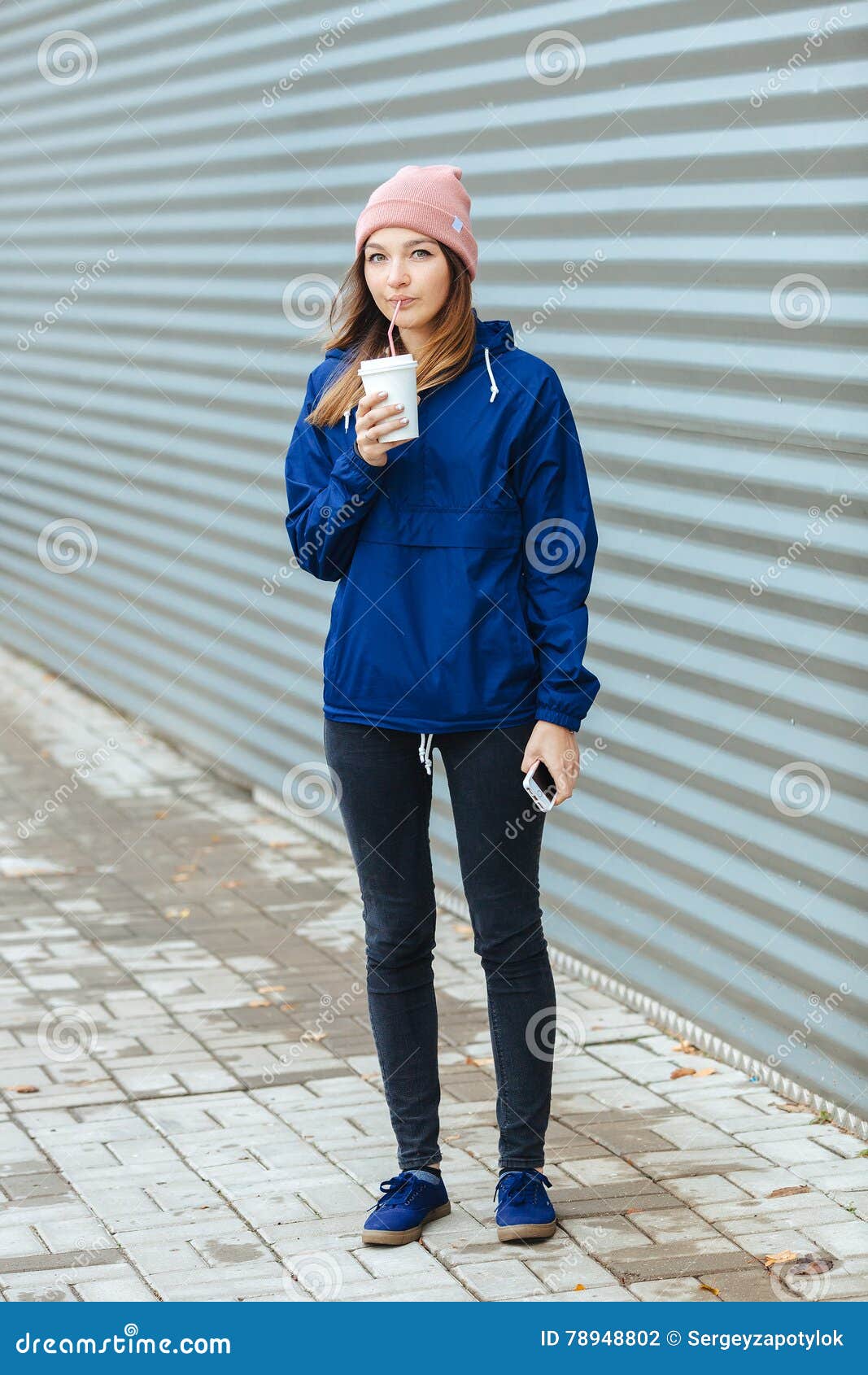 stylish sporty brunette woman in trendy urban outwear posing with big white disposable cup straw cold rainy fall day against strip